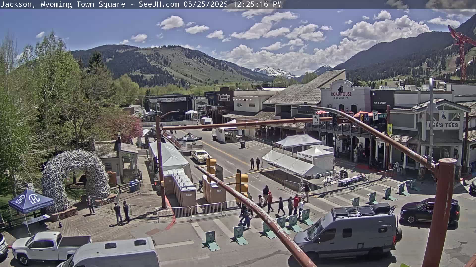 A town square with buildings, vehicles, and people under a partly cloudy sky.