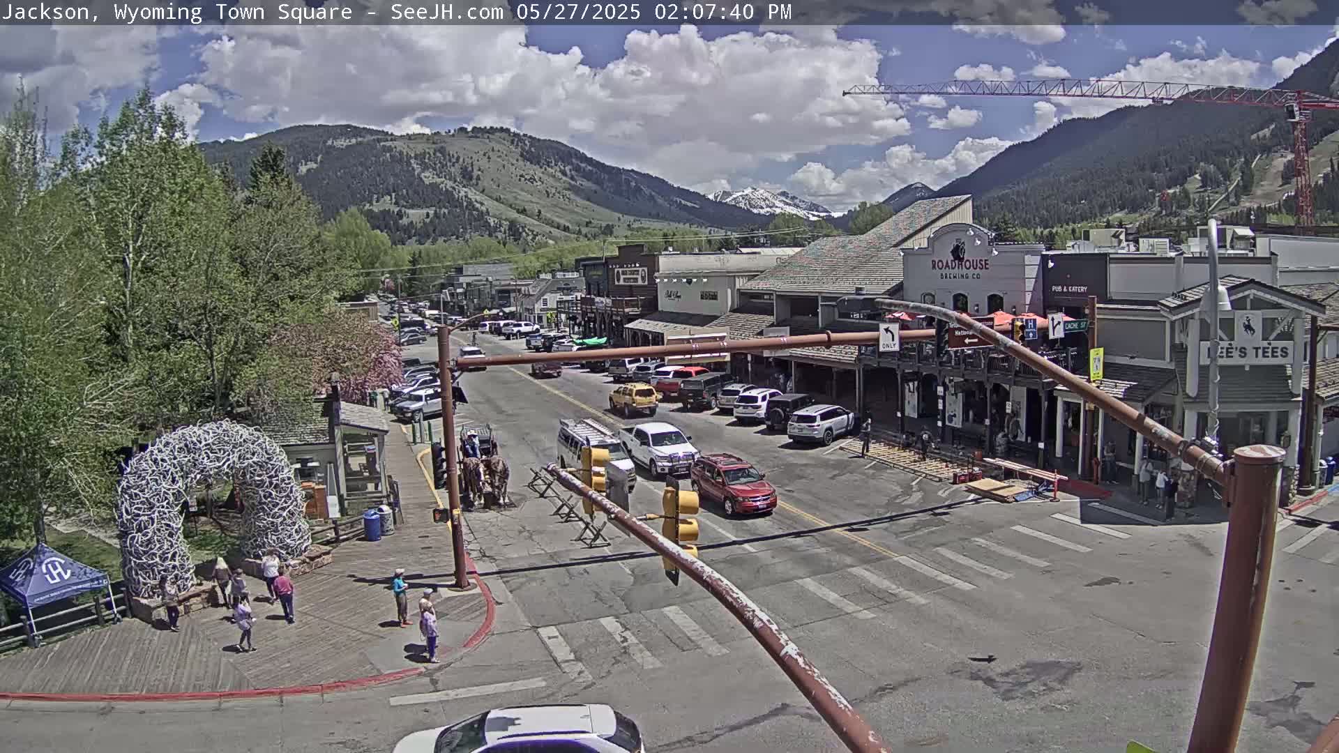 A sunny town square, filled with cars and pedestrians, sits nestled amongst green trees and mountains under a partly cloudy sky.
