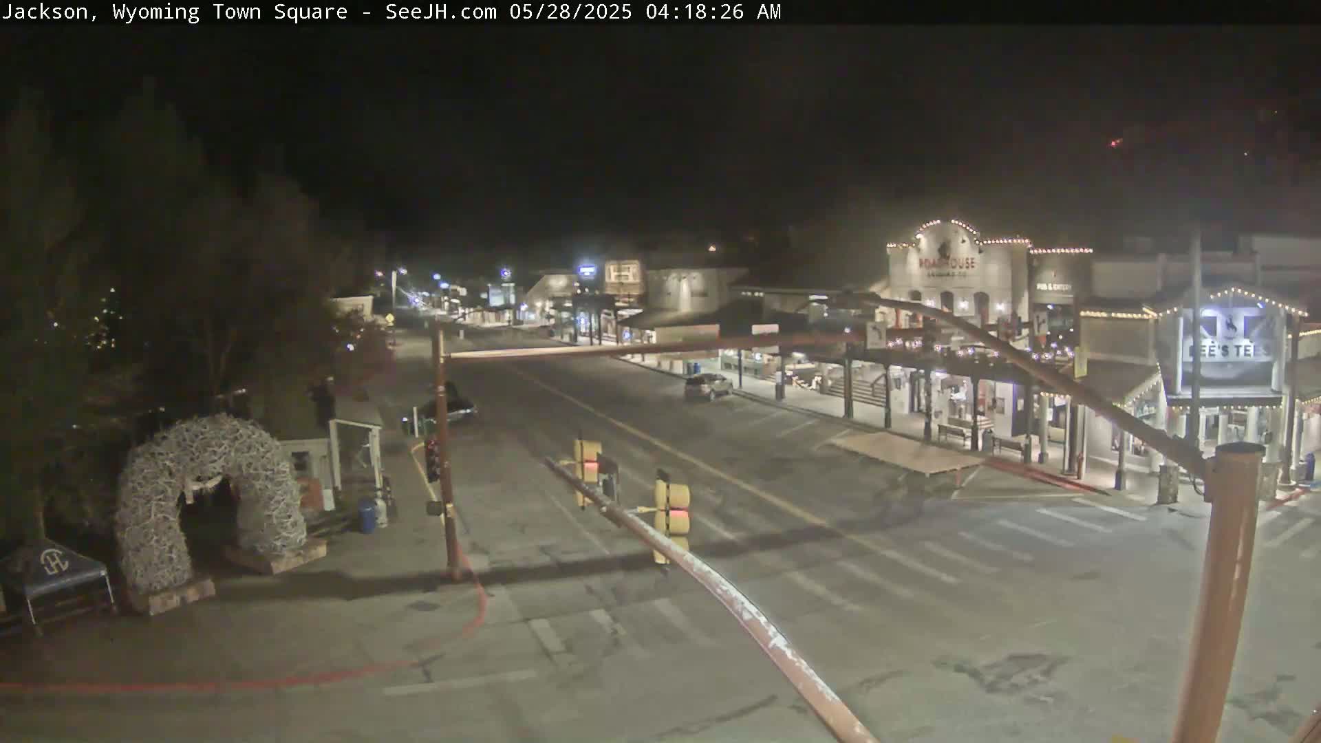A mostly empty town square at night, illuminated by streetlights and building lights, features a large antler archway and several buildings along the street.