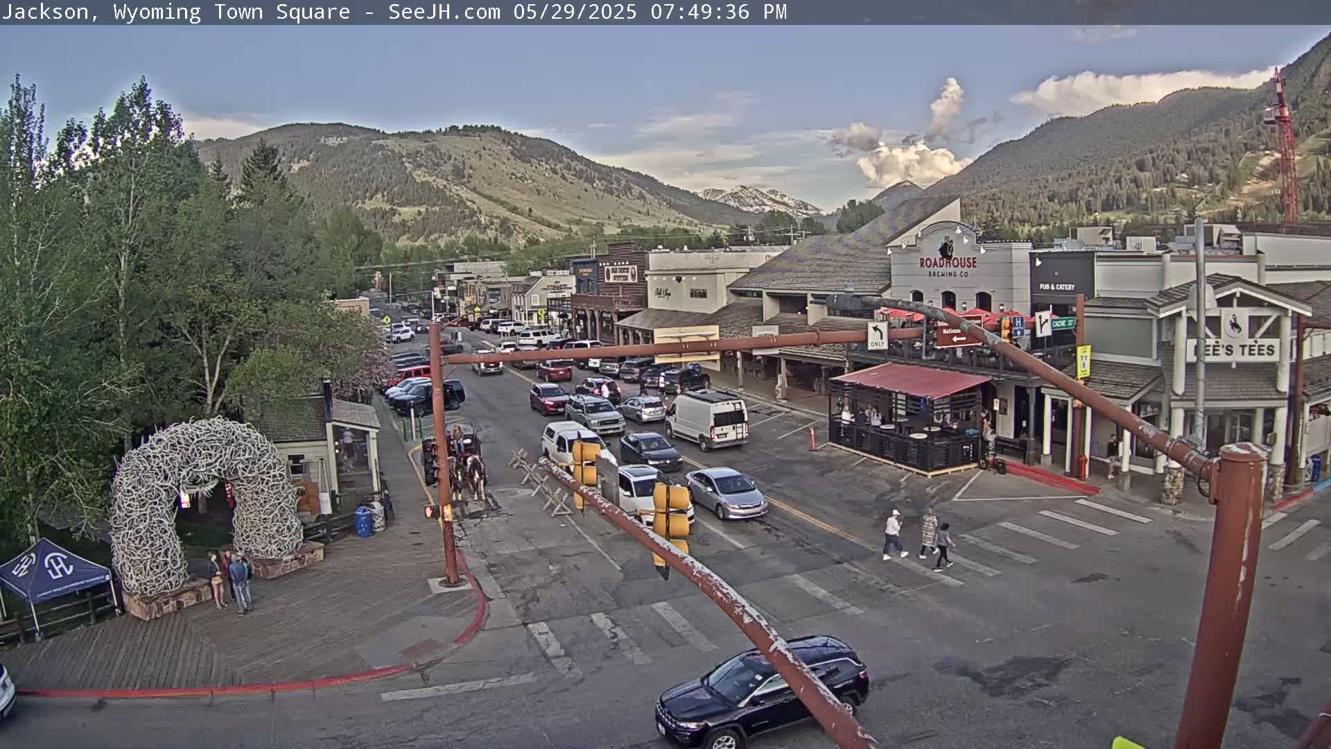 A partly cloudy afternoon view of a town square with vehicles on the road, buildings lining the street, and a large antler archway in the foreground.