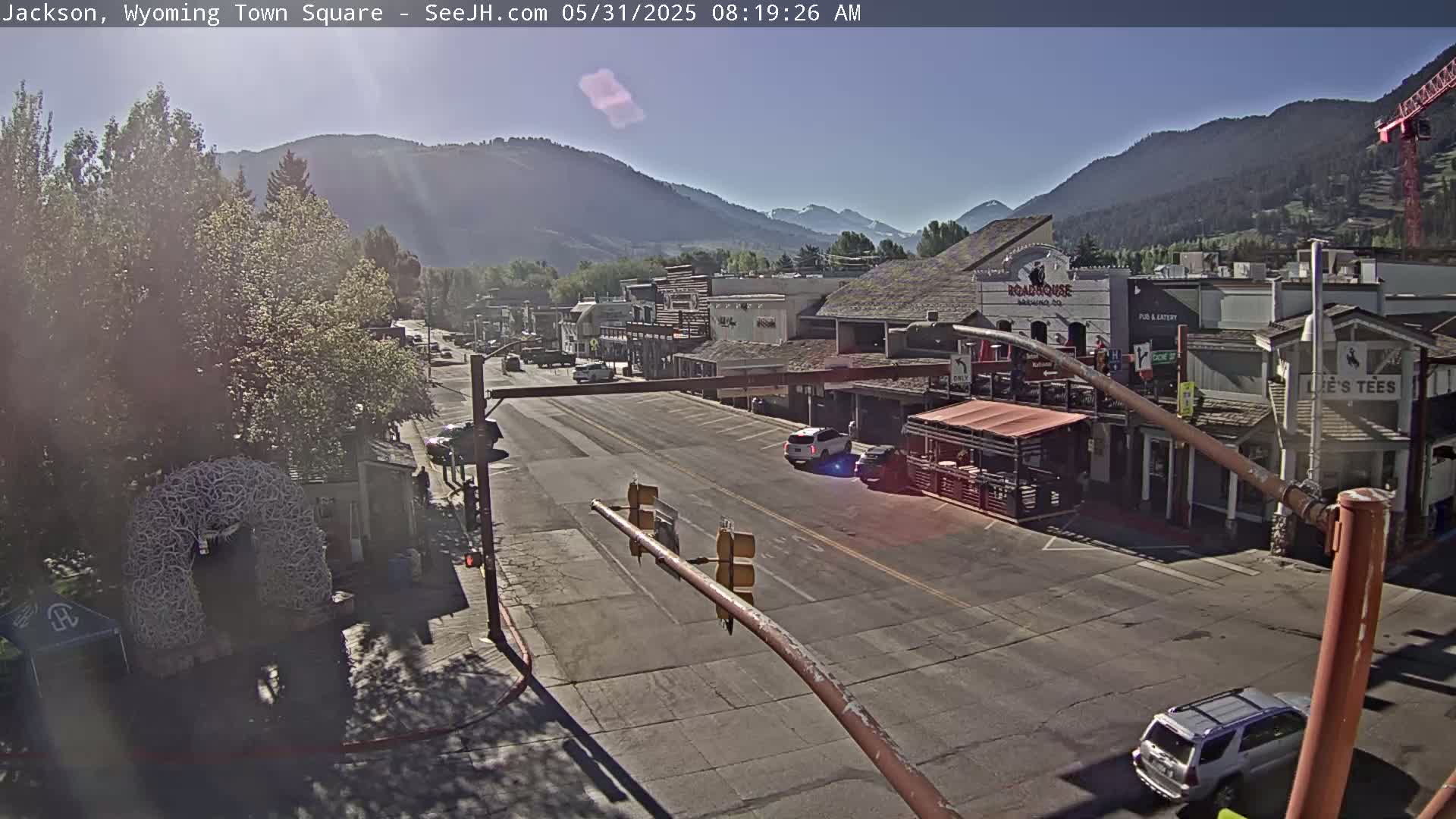 A sunny morning view of a town square with several buildings, a street with cars, and mountains in the background.