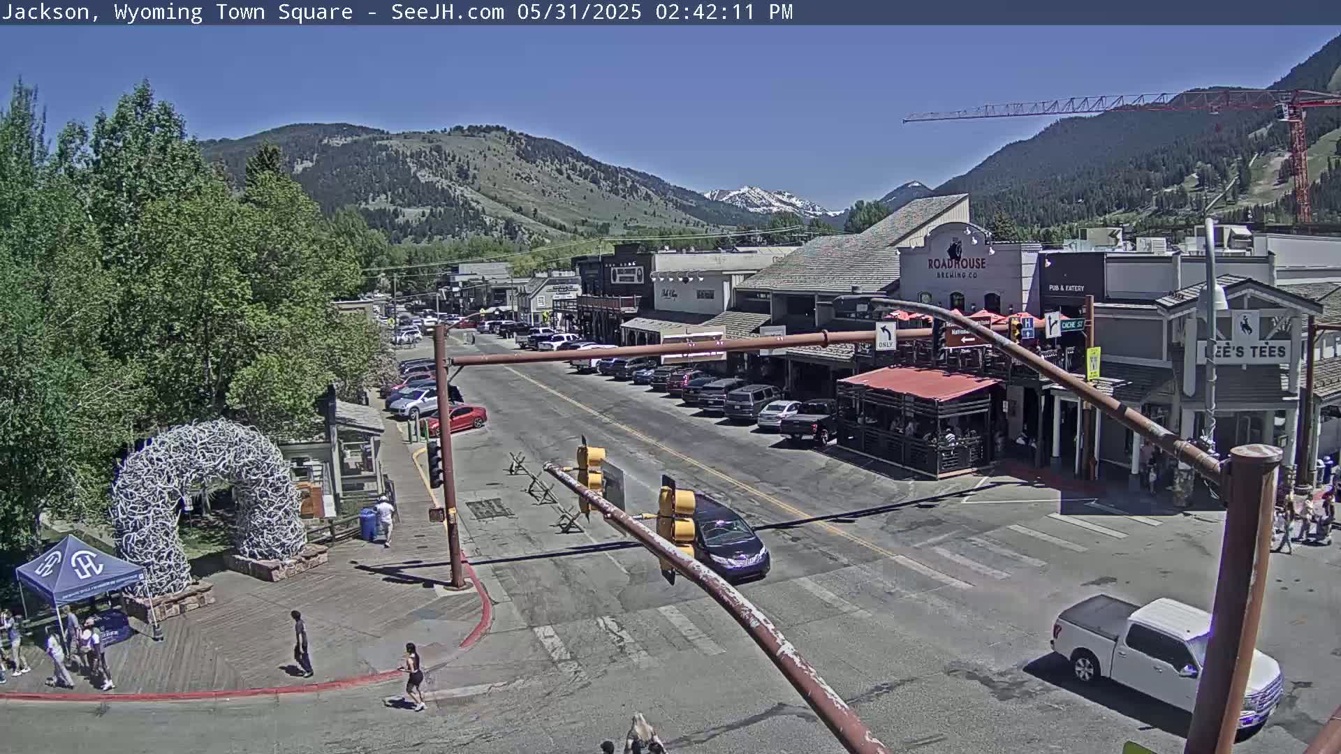 A town square scene on a sunny day shows a street with parked cars and buildings, a large antler arch, and a few pedestrians.