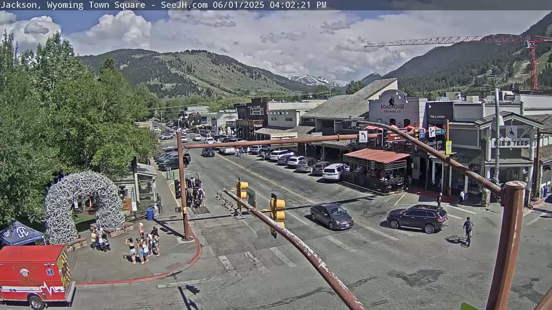 A partly cloudy day in a town square, with cars parked along the street and people gathered near a large antler arch, with mountains in the background.