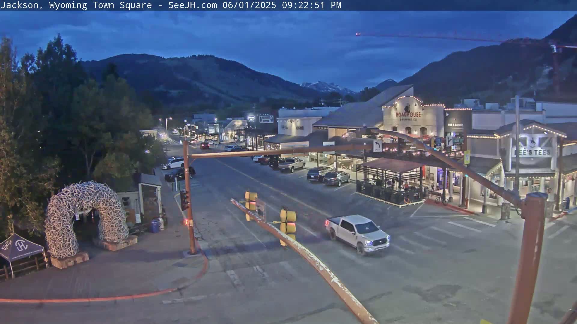 A town square at dusk, with cars on the street in front of shops and restaurants, under a mostly clear sky.