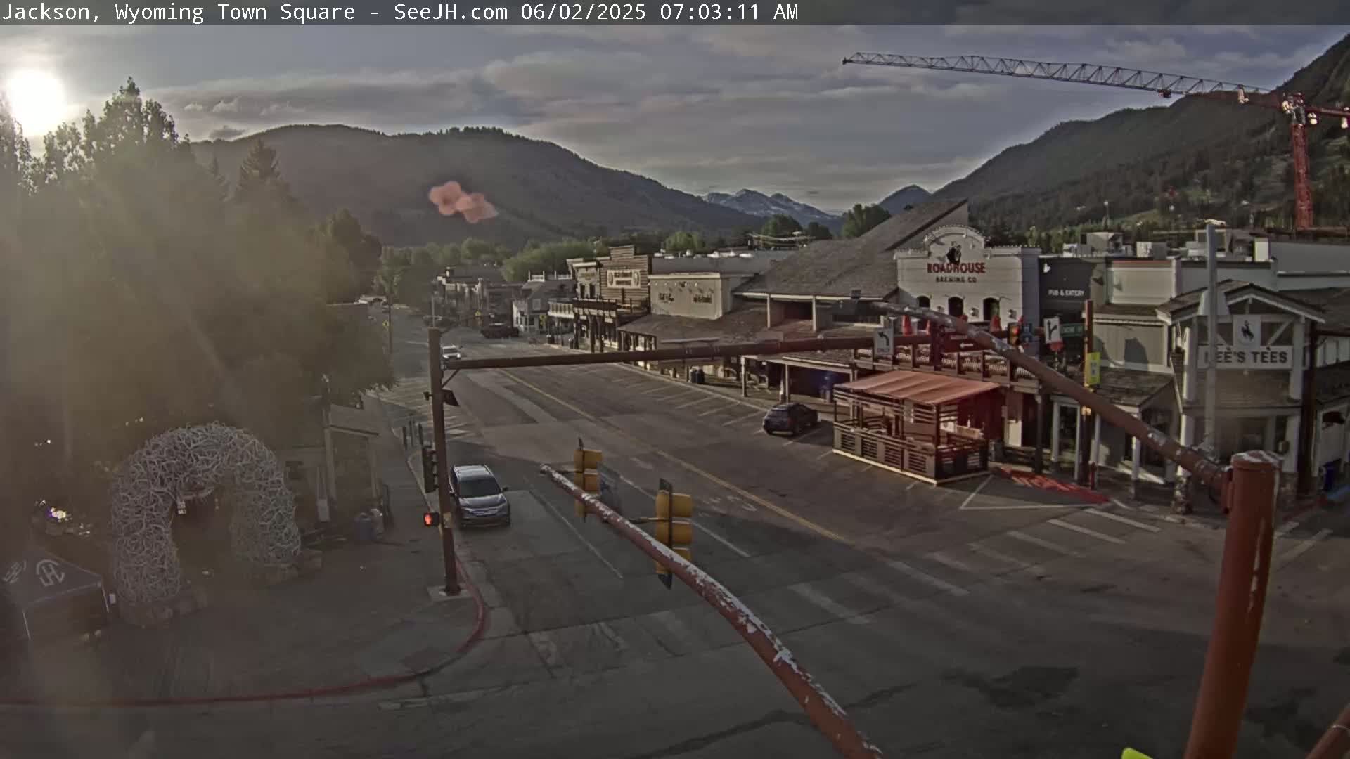 A mostly sunny morning scene shows a town square with a few cars, some buildings, and mountains in the background.