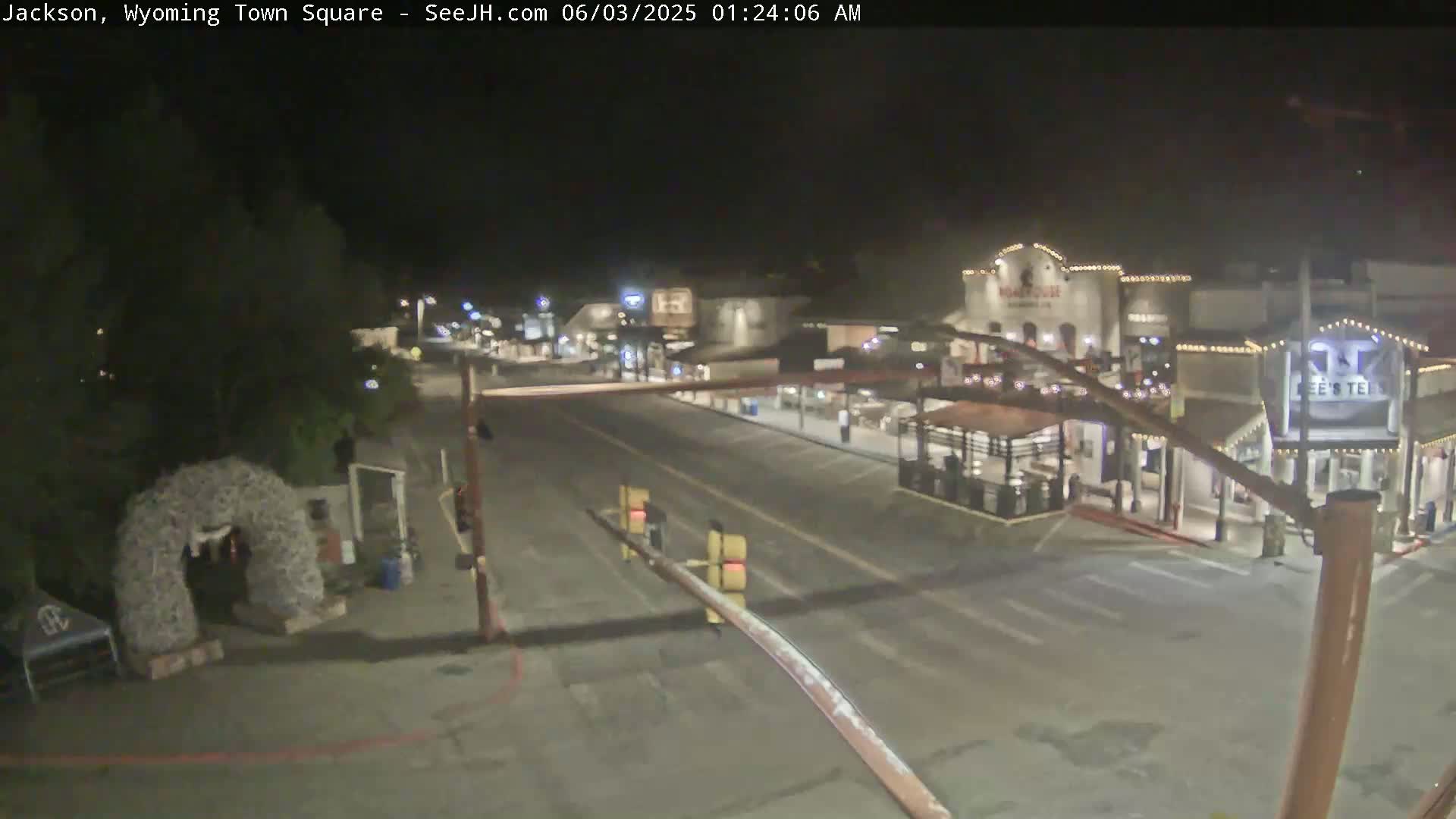 A mostly empty town square at night, with a few buildings illuminated and some vehicles parked along the street.