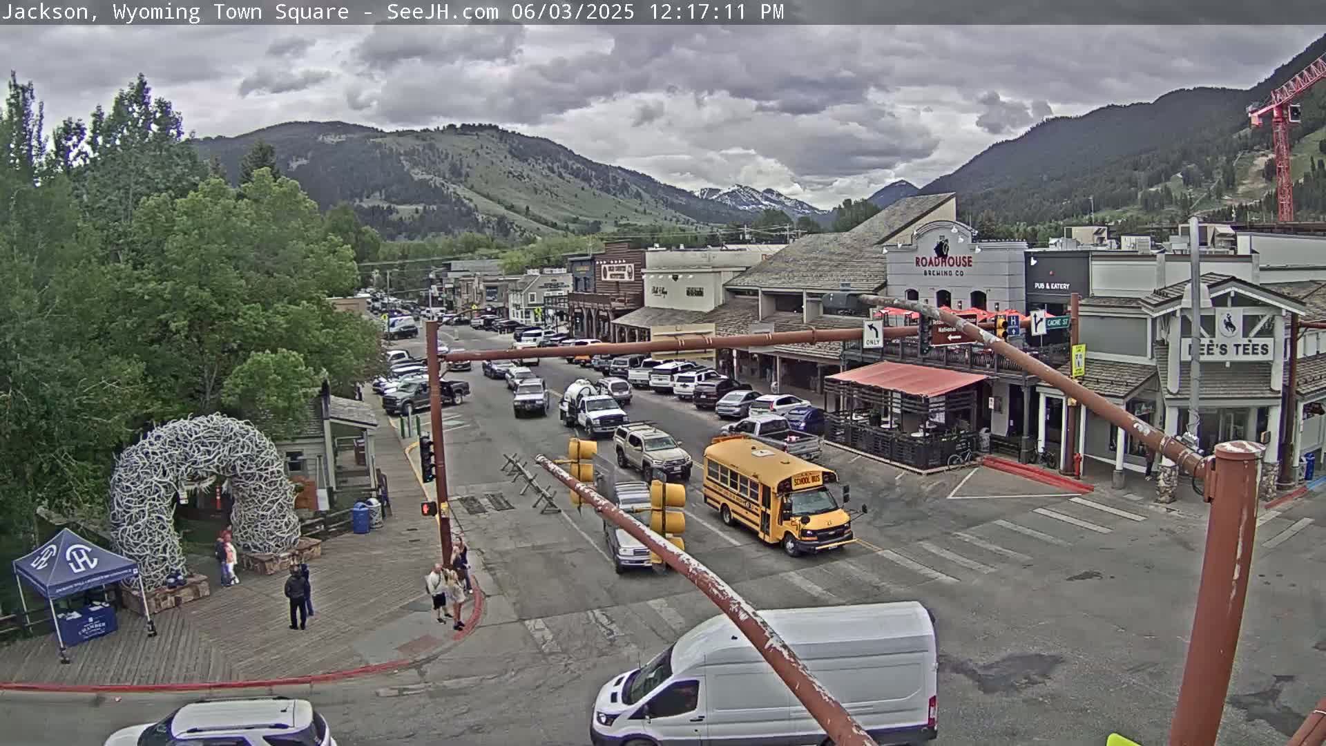A town square with parked and moving cars under a partly cloudy sky, backed by mountains.