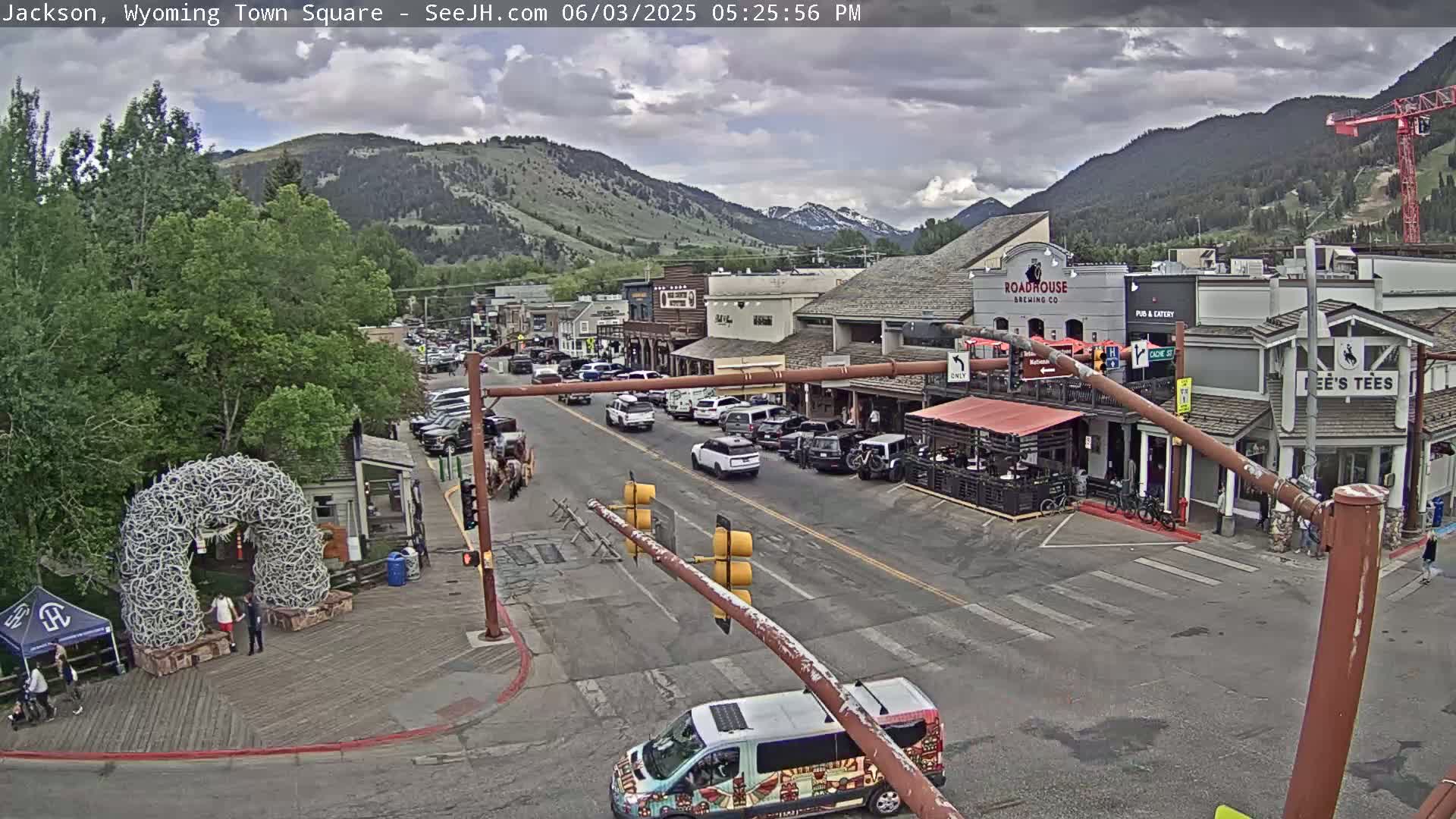 A partly cloudy day shows a town square with several buildings, vehicles, and pedestrians, set against a backdrop of mountains.