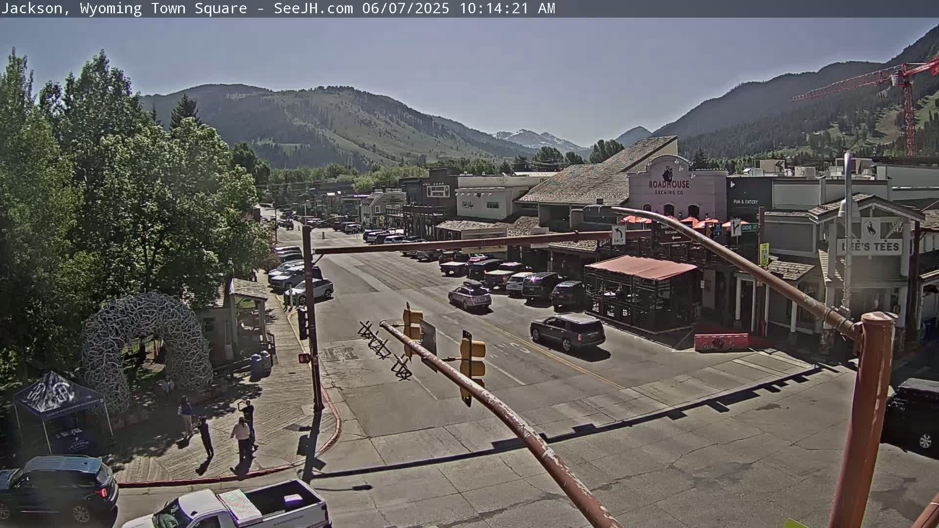 A sunny town square scene shows a street with cars, buildings, and people, nestled in a valley surrounded by mountains.