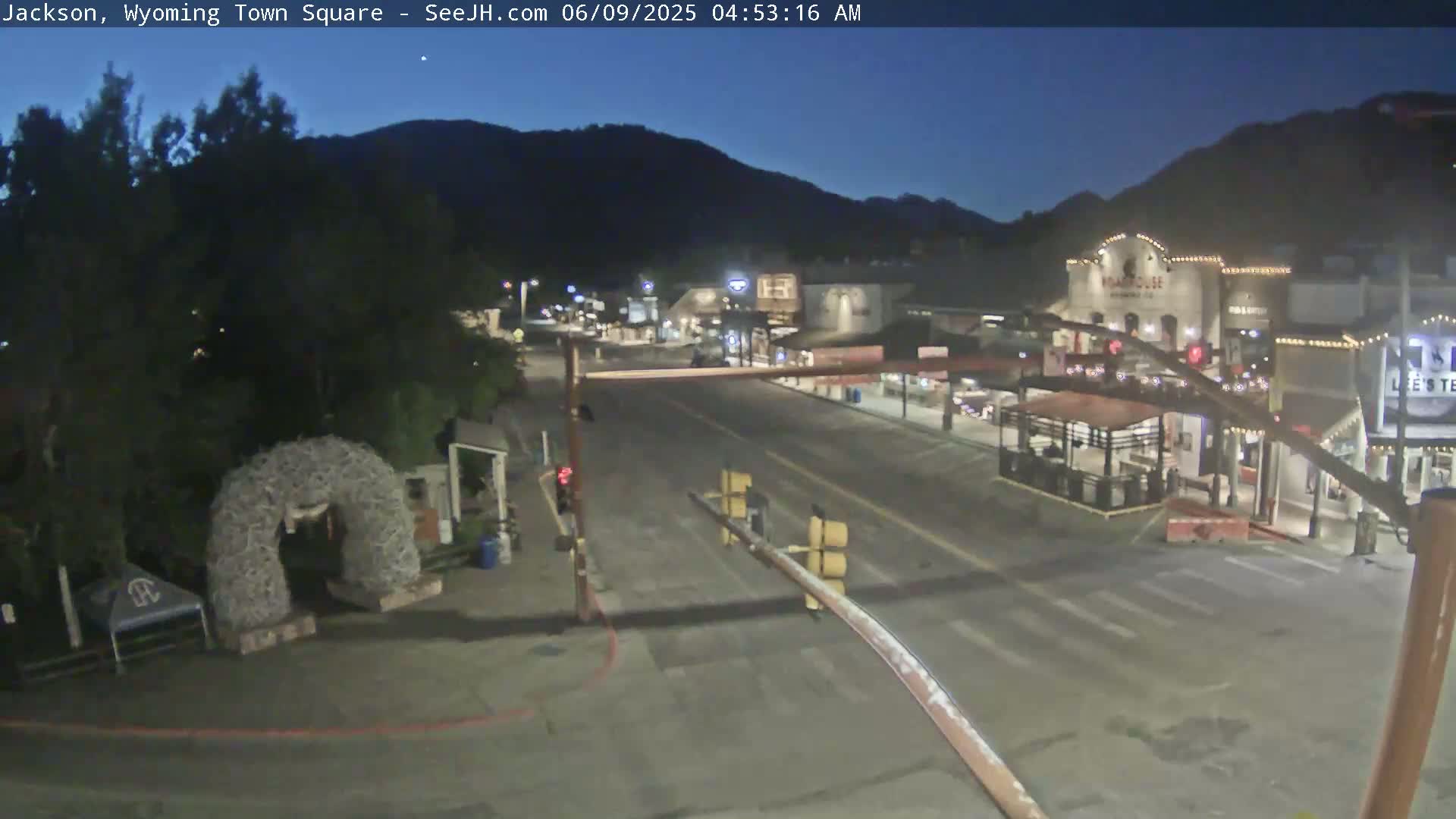 A mostly empty town square at night, under a dark, clear sky, is bordered by buildings with some lights on, and a large, dark mountain is visible in the background.
