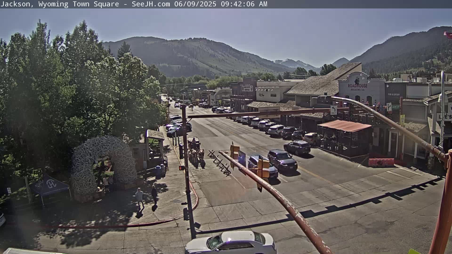 A town square with parked cars, buildings, and a horse-drawn carriage on a sunny day in front of mountains.