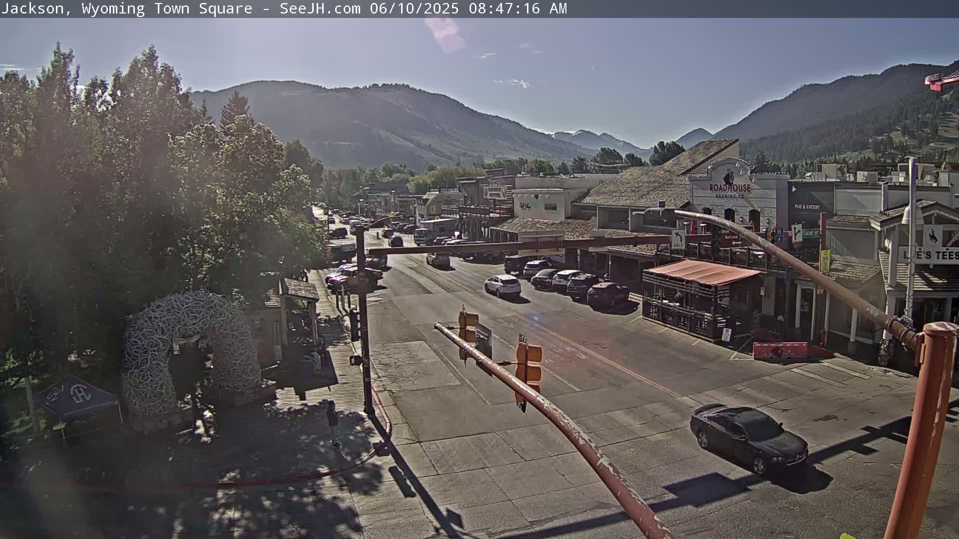 A sunny day reveals a town square with a street lined with parked cars and buildings, nestled against a backdrop of mountains.