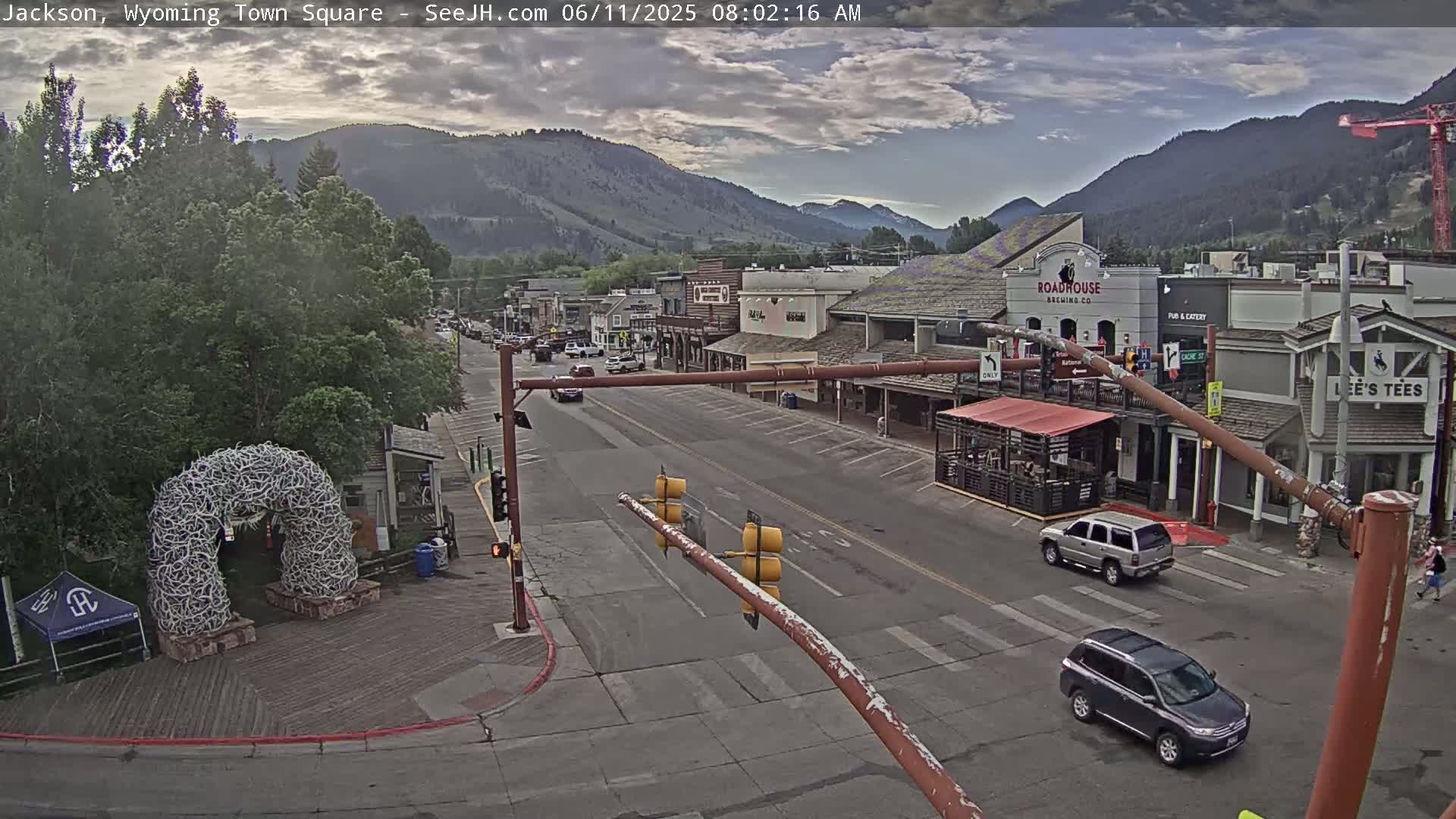 A mostly cloudy morning shows a town square with several buildings, a few cars on the road, and an antler archway in the foreground.