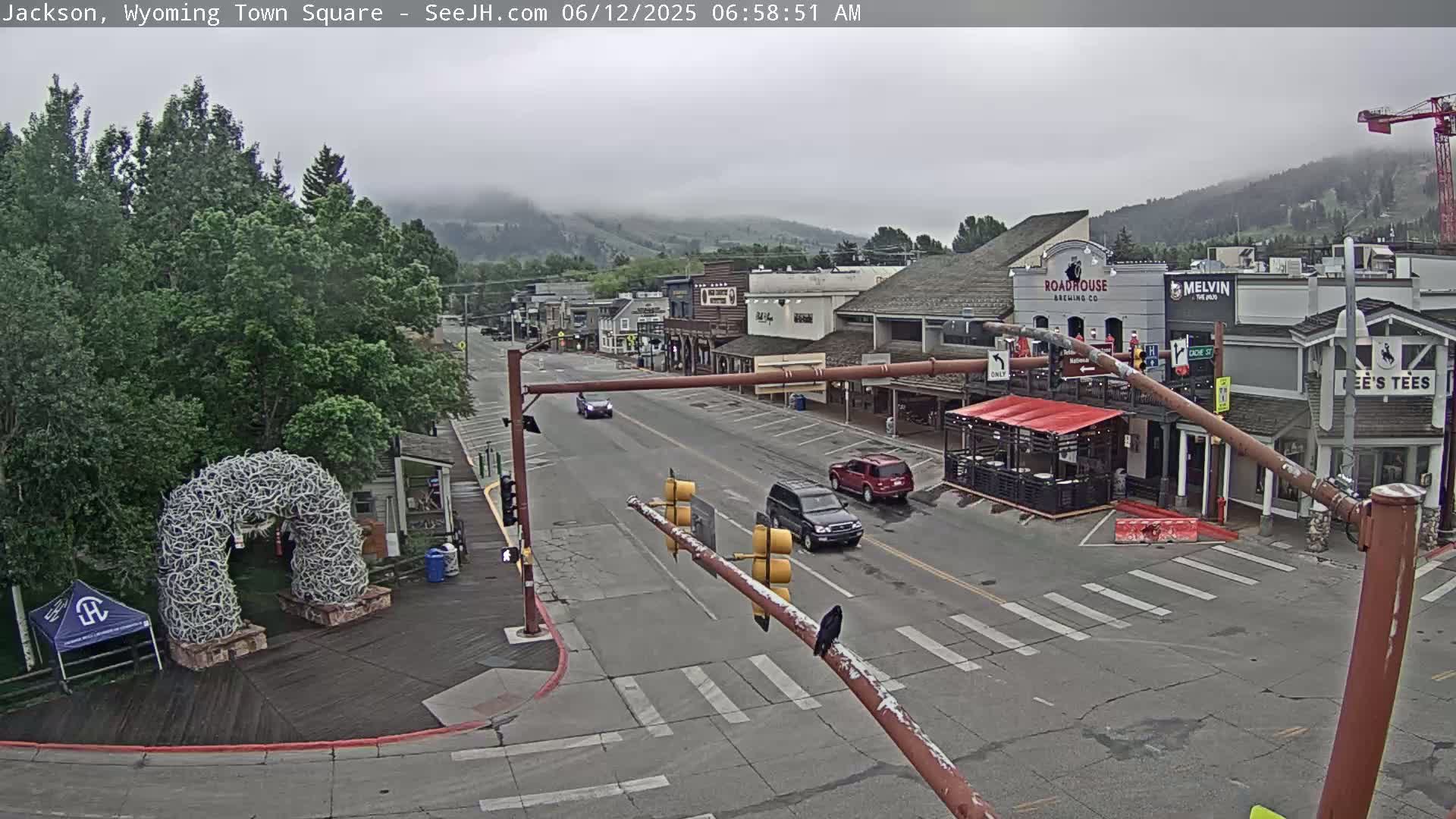 A mostly cloudy morning scene shows a town square with several buildings, cars on a street, and a large antler arch.