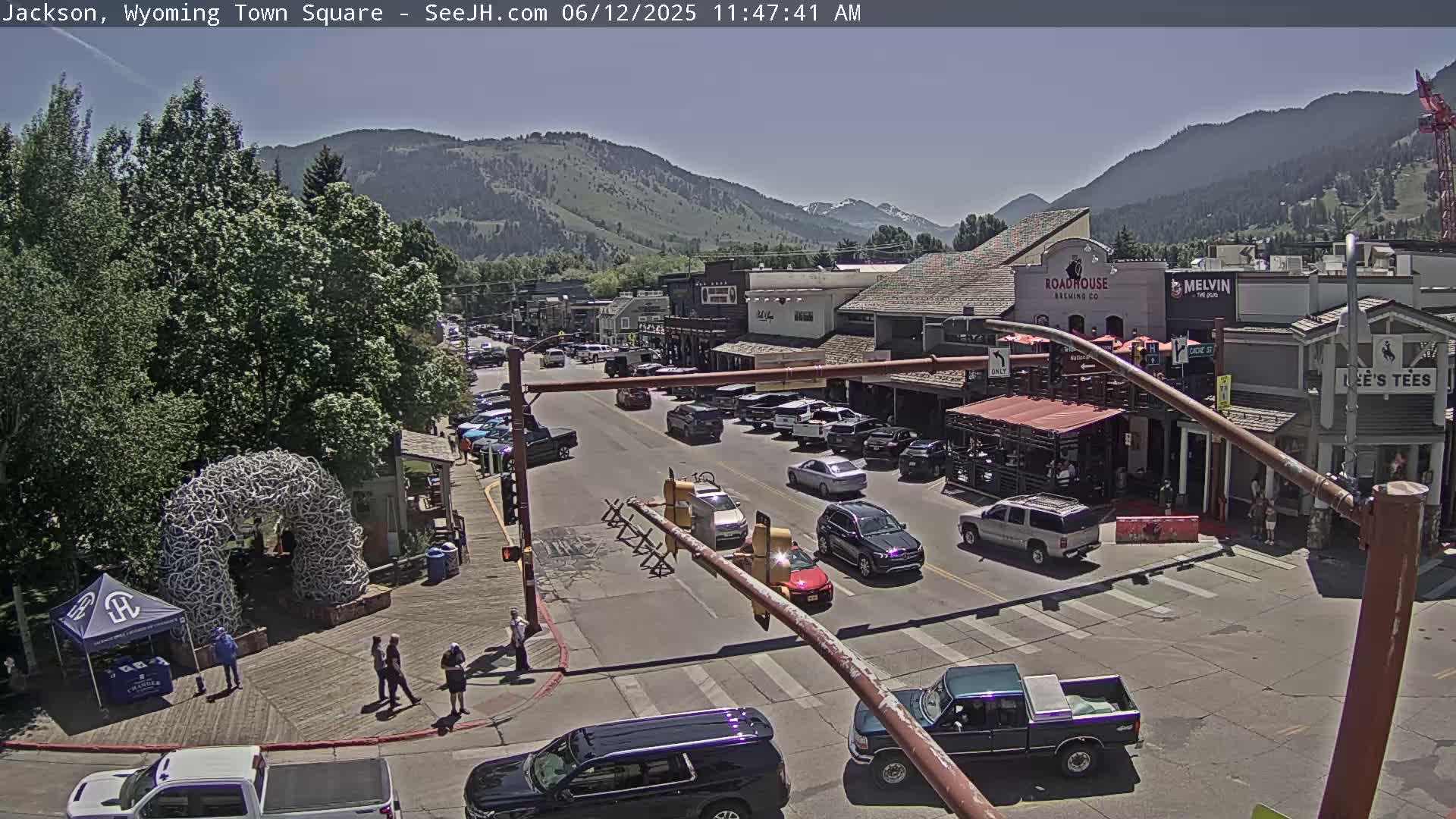 A sunny day in a town square shows cars driving on a street lined with buildings, and pedestrians walking on a sidewalk next to a large antler arch.