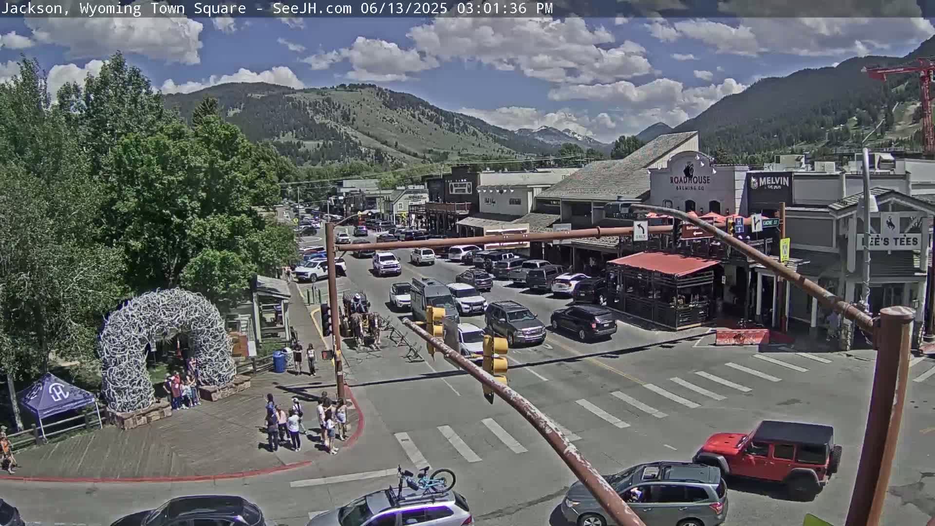 A town square with a crowd of people, numerous vehicles, and buildings under a partly cloudy sky, set against a backdrop of mountains.