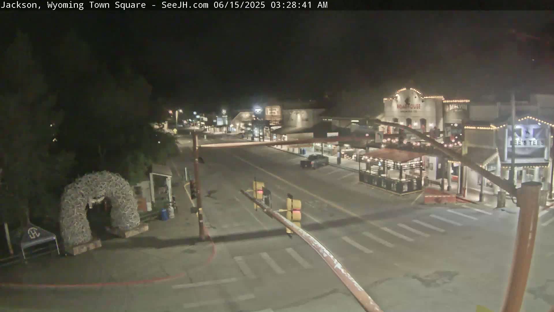 A nighttime, mostly empty town square with a few vehicles, several buildings with lights on, and a large antler archway.