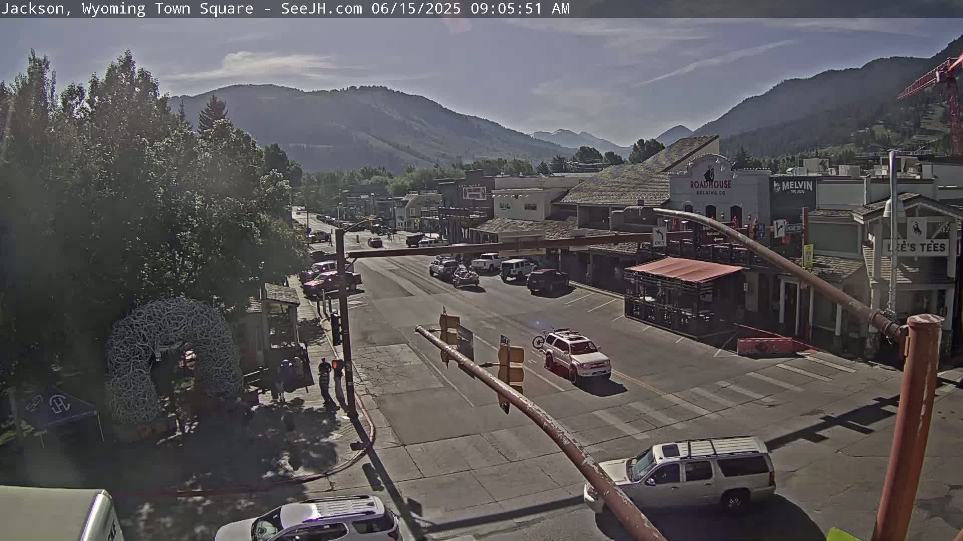 A mostly sunny view of a town square, with cars parked and driving on a street lined with buildings, and mountains in the background.