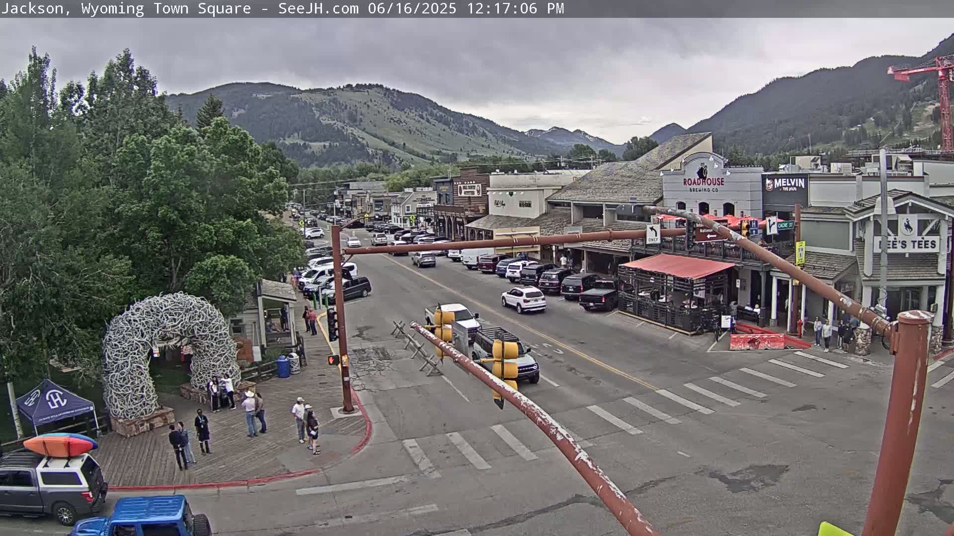 A town square with parked cars and pedestrians, under an overcast sky, is situated against a backdrop of mountains.