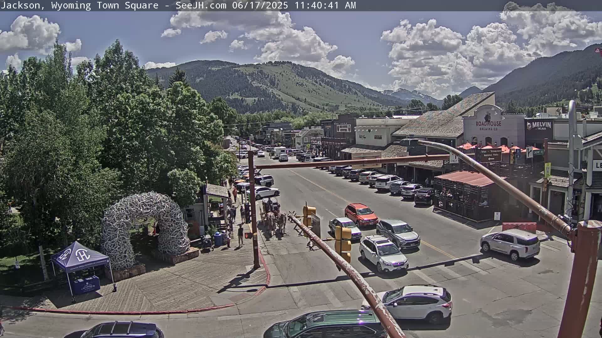 A town square with parked cars and a horse-drawn carriage on a sunny day with partly cloudy skies and mountains in the background.