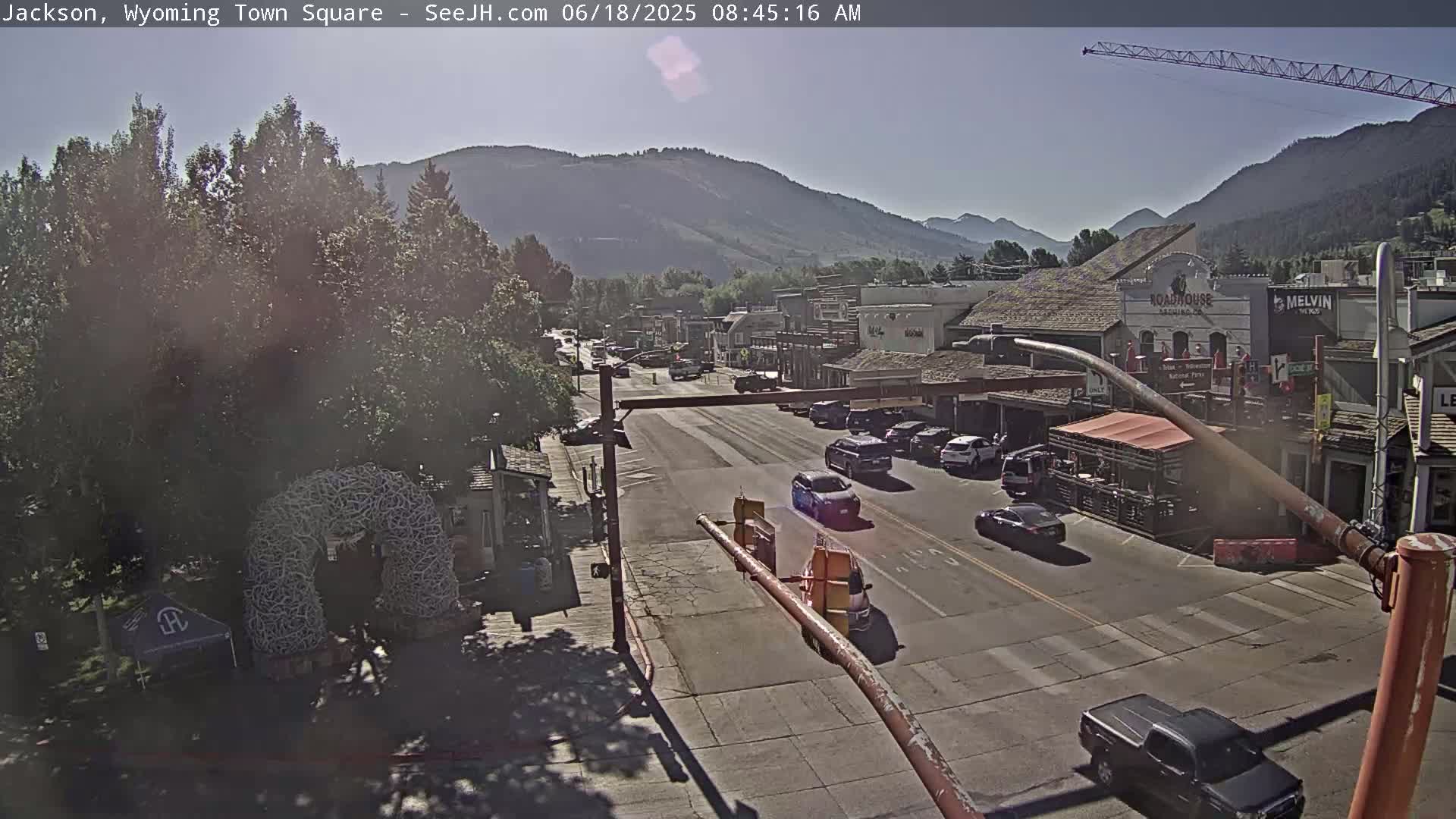 A sunny day reveals a town square with a main road flanked by buildings, mountains in the background, and several cars driving through.