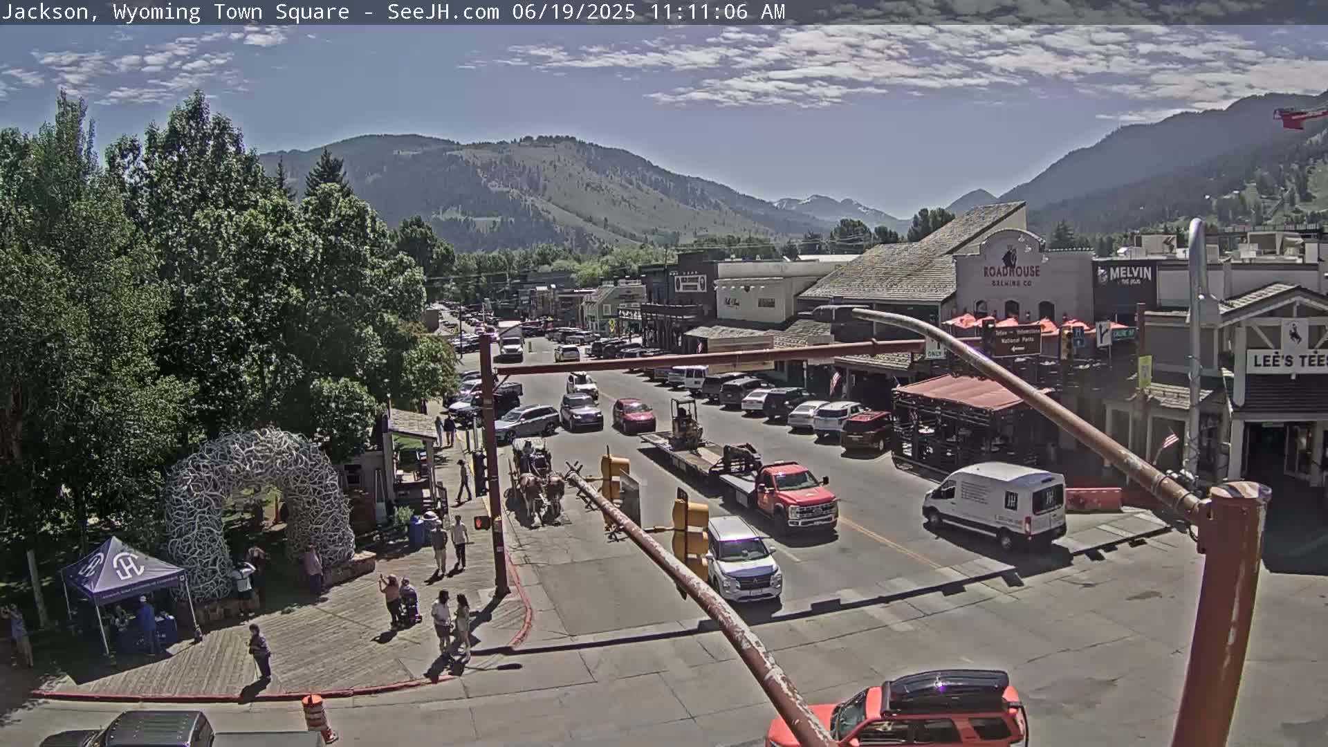A town square with parked cars and pedestrians, under a partly cloudy sky, is situated at the base of a mountain range.