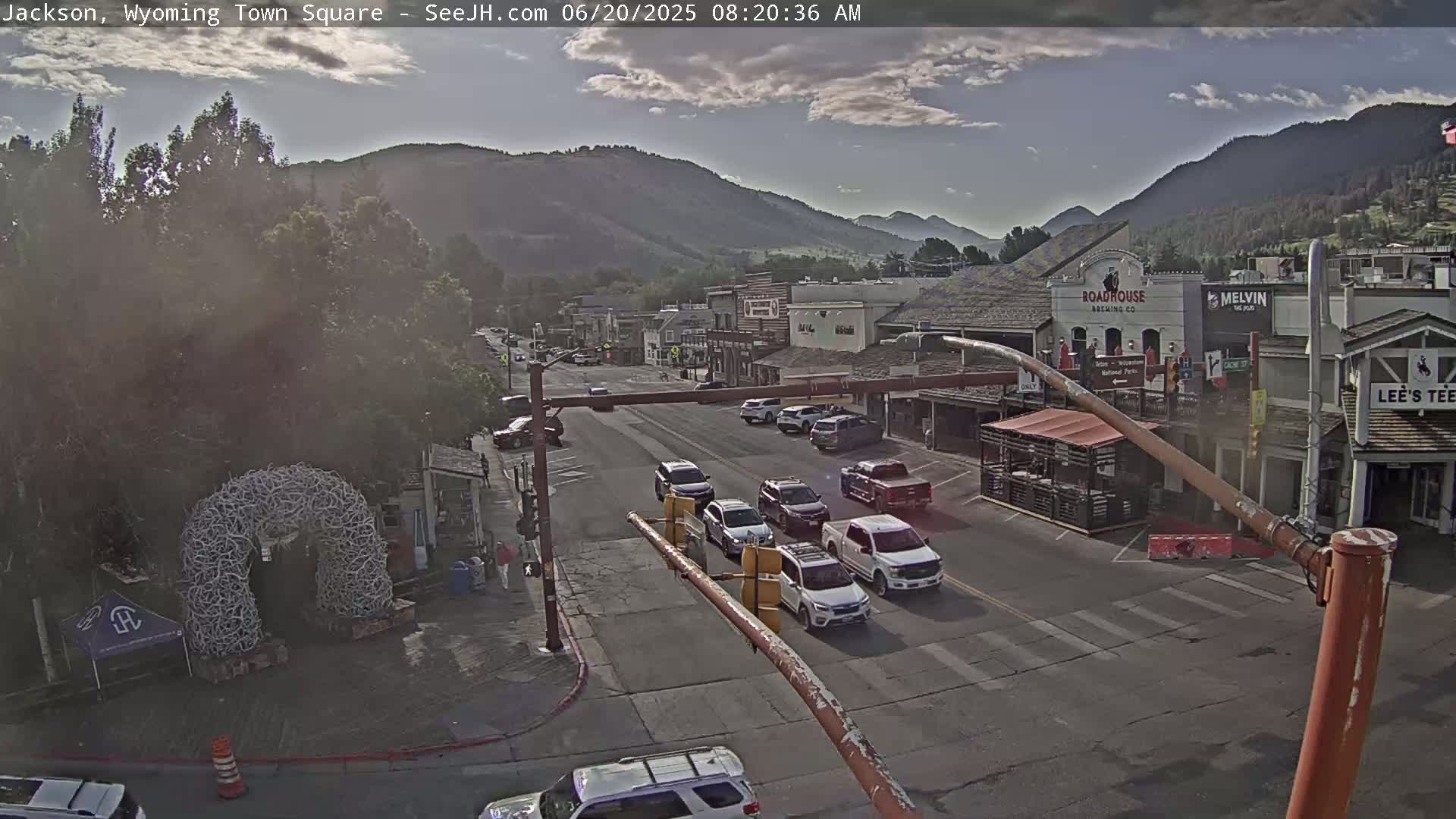 A partly cloudy morning scene shows a town square with several vehicles driving on a street lined with buildings and surrounded by mountains.