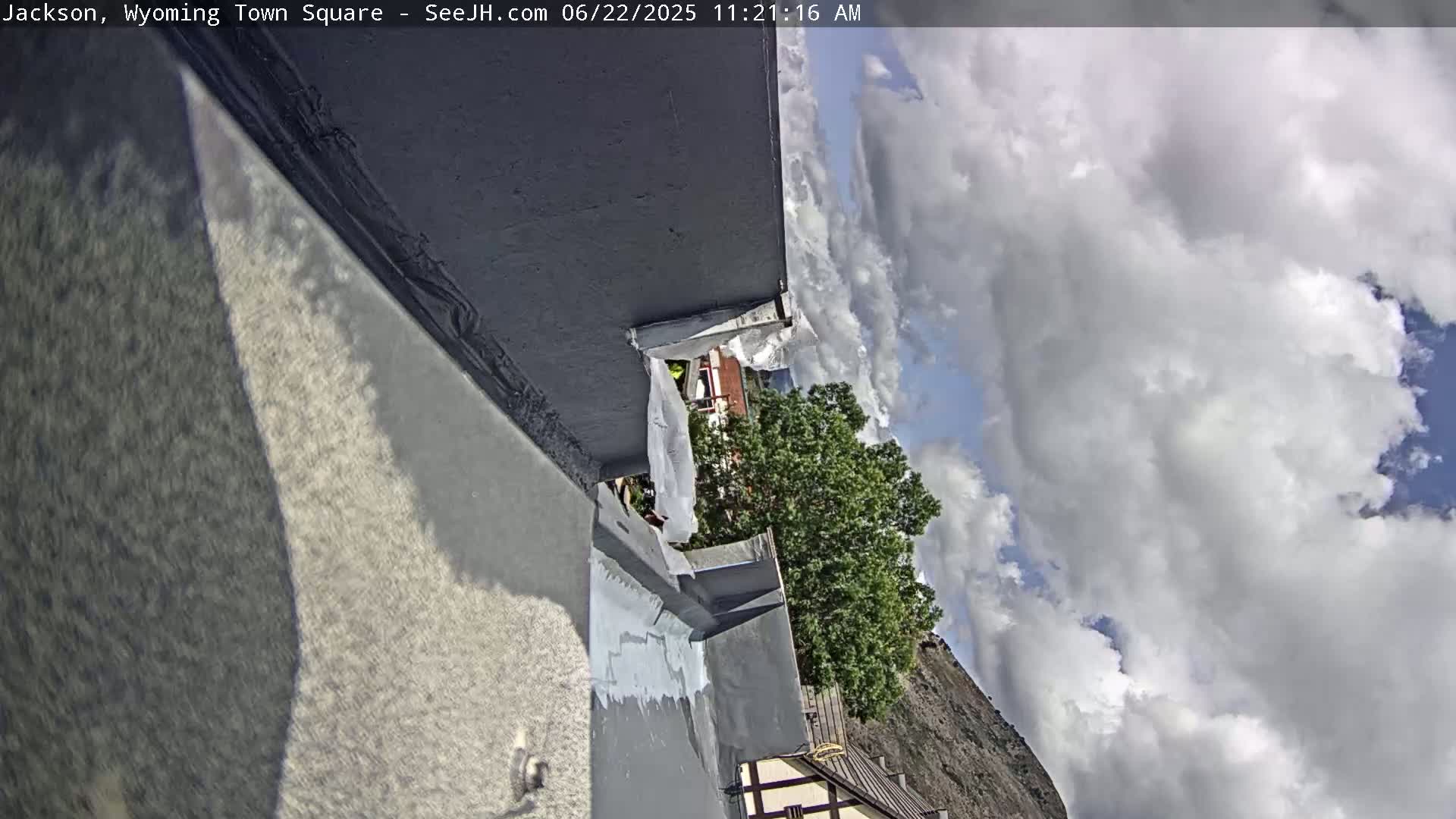 A partially cloudy sky is visible above a gray, flat rooftop undergoing repairs, with a glimpse of green trees and a building in the background.