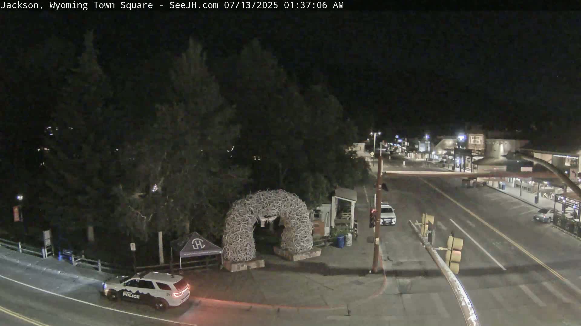 A nighttime view of a town square, featuring a large antler archway, a police car, and a sparsely populated street.