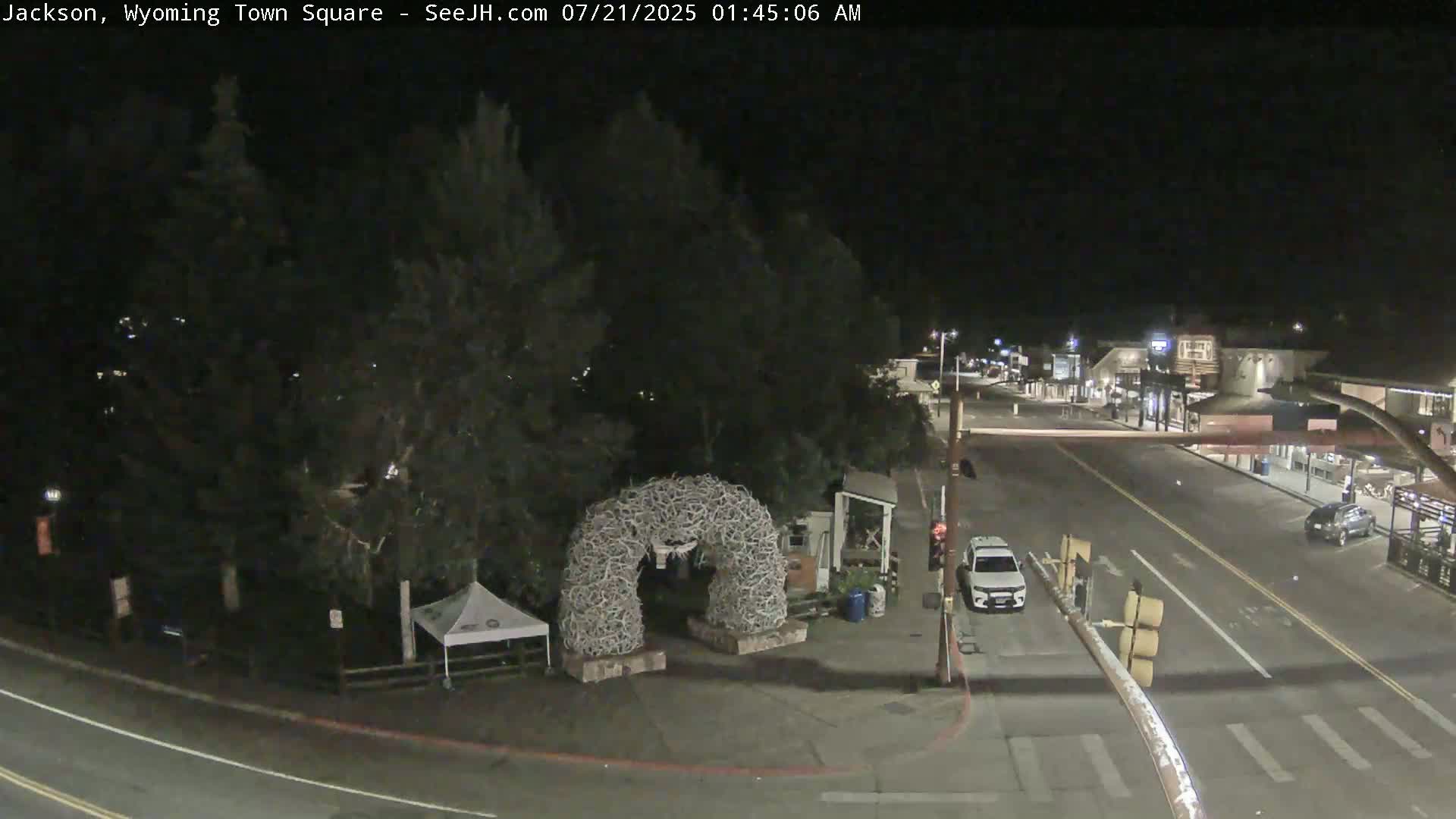 A nighttime view of a town square with a large antler arch, a few vehicles, and some buildings visible in the distance under a clear, dark sky.