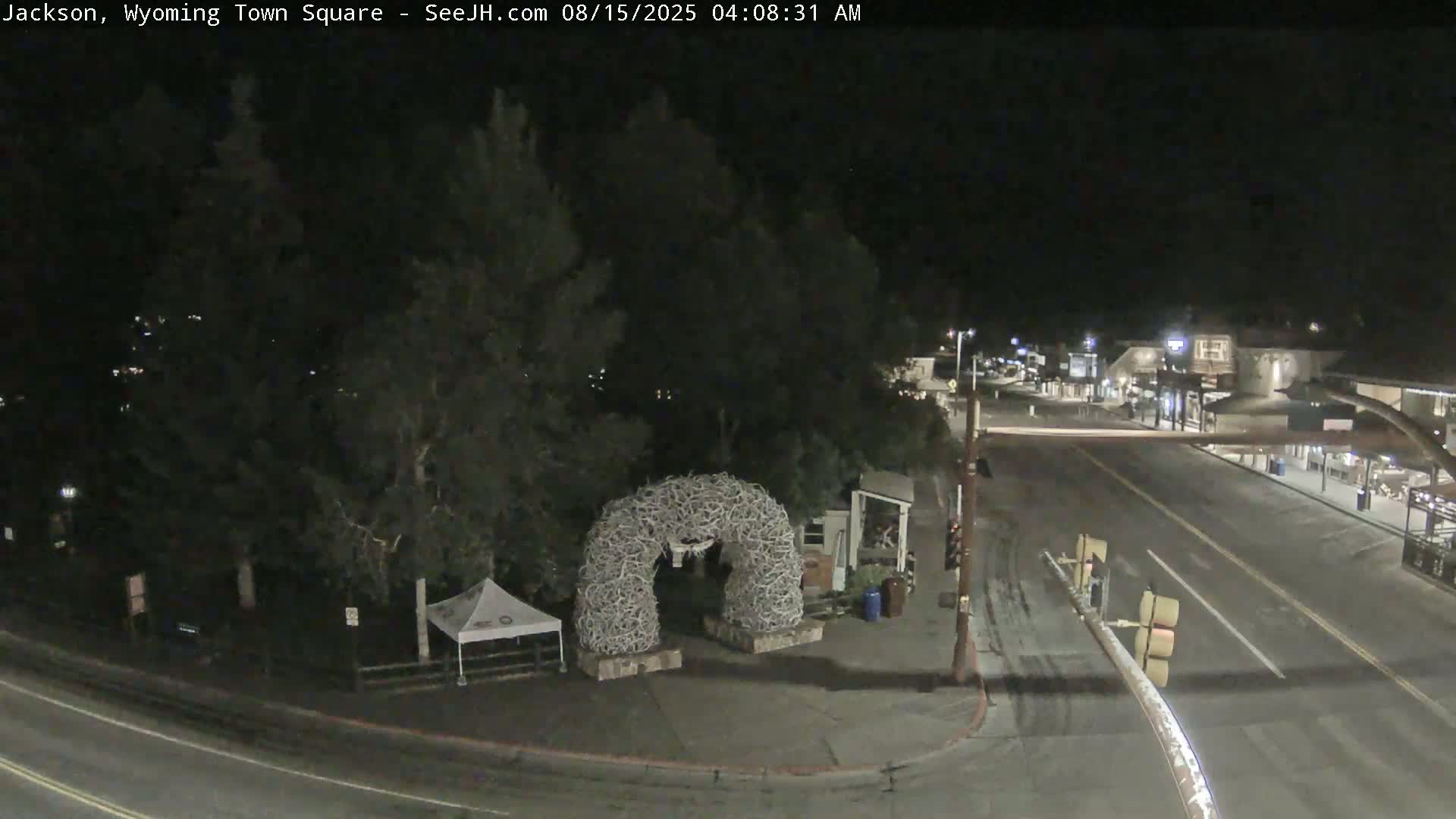 A nighttime view of a town square with a large antler arch, a few vehicles, and some buildings visible in the distance under a clear, dark sky.