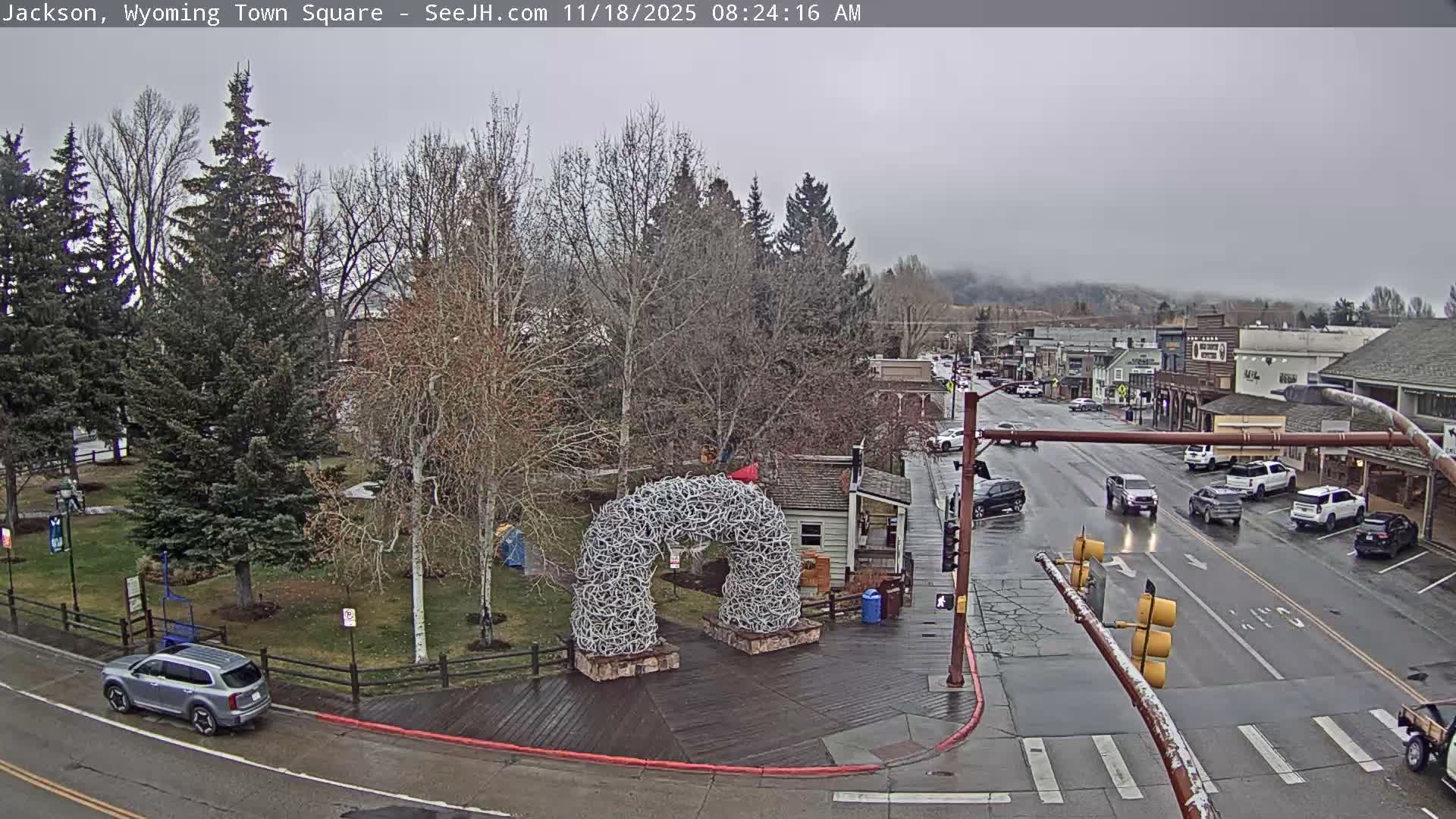 The wet streets of a town square reflect the lights on a dark, rainy night, featuring a prominent antler arch, bare trees, and surrounding buildings.