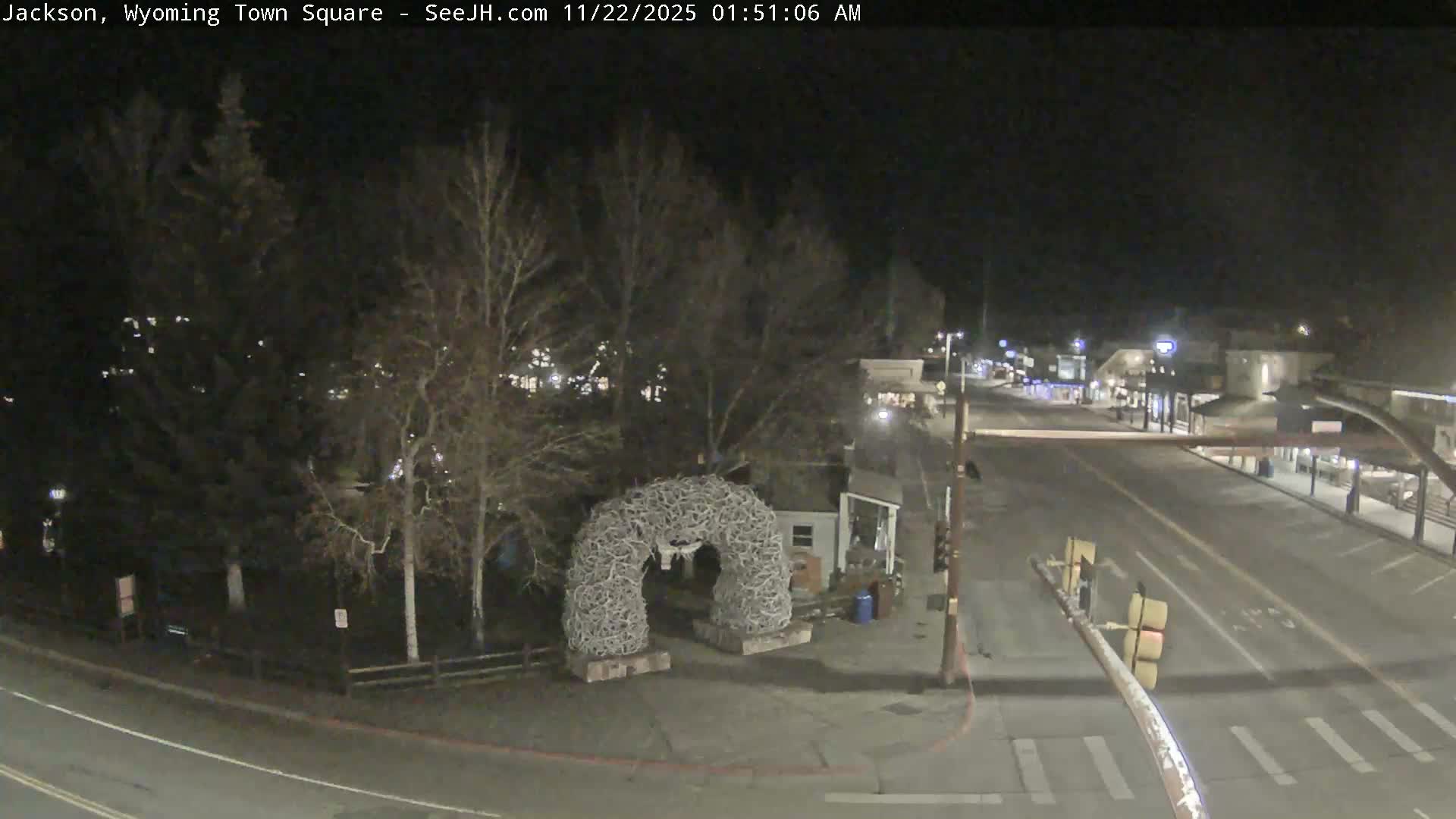 On a clear, dark night, a town square is visible with a large antler archway, bare trees, and illuminated buildings along a street with a red traffic light.