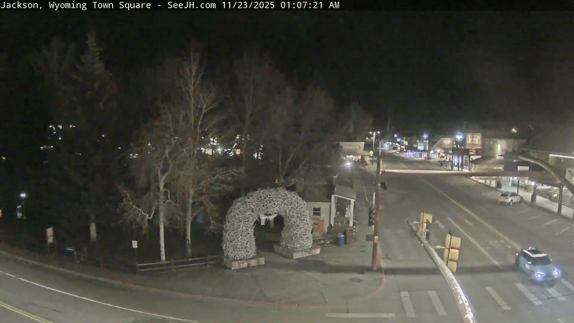 A clear night provides an overhead view of a brightly lit town square, featuring a large antler archway and bare trees alongside a street with faint vehicle light trails and illuminated buildings.