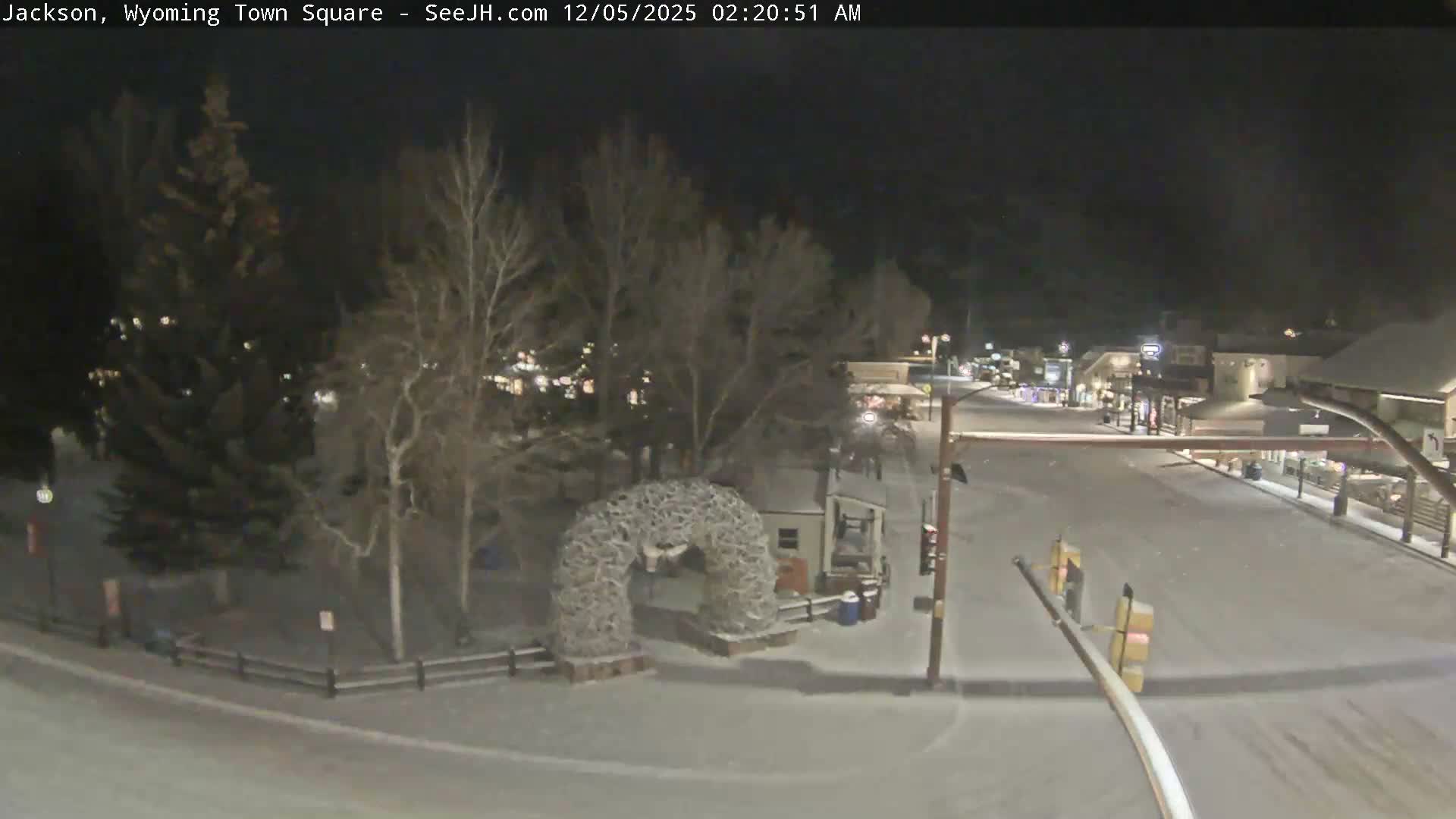 A snow-covered street and park area featuring a prominent antler archway are illuminated by artificial lights on a dark, snowy night, flanked by bare trees and buildings.
