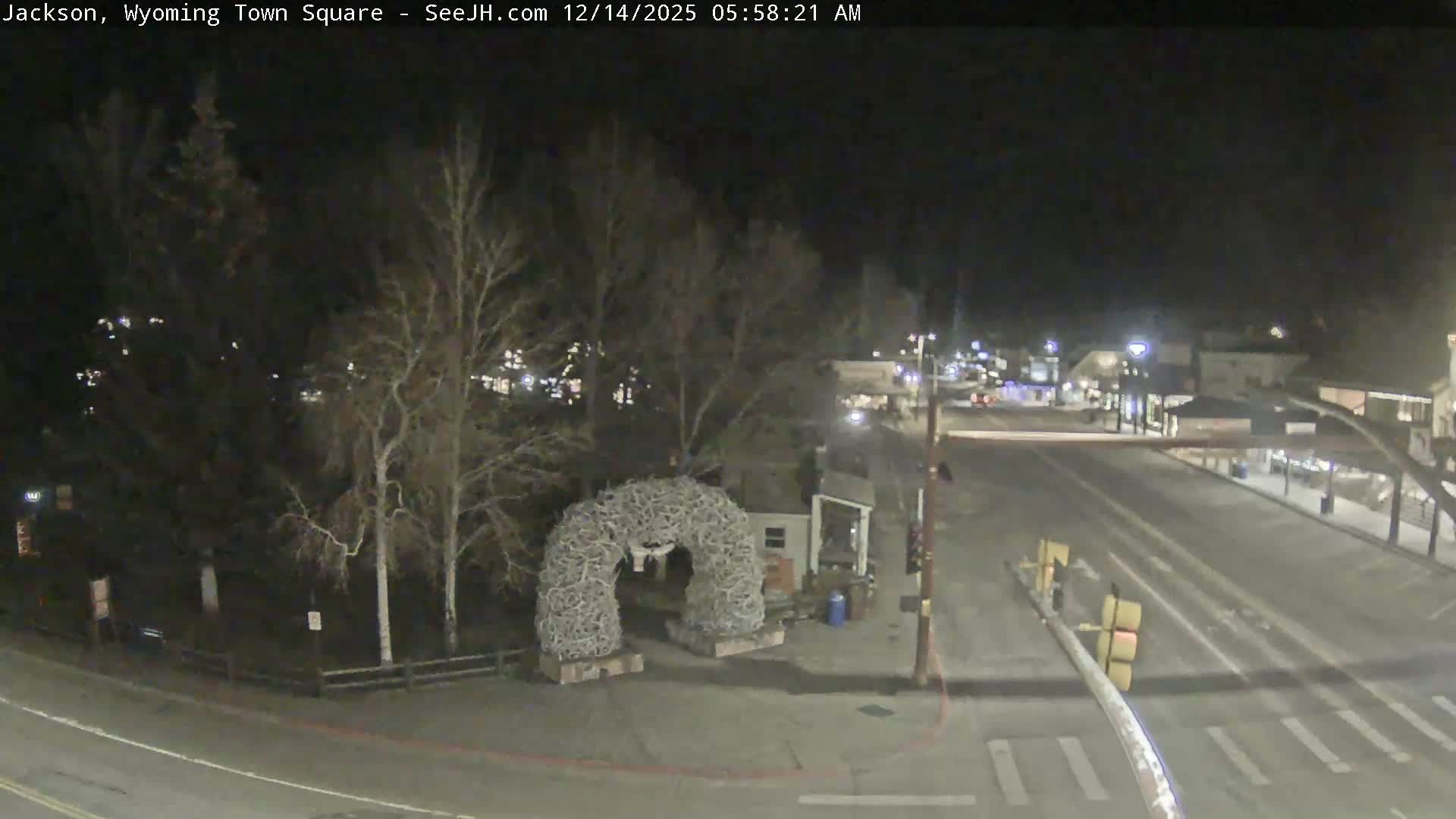 A snow-covered street and park area featuring a prominent antler archway are illuminated by artificial lights on a dark, snowy night, flanked by bare trees and buildings.