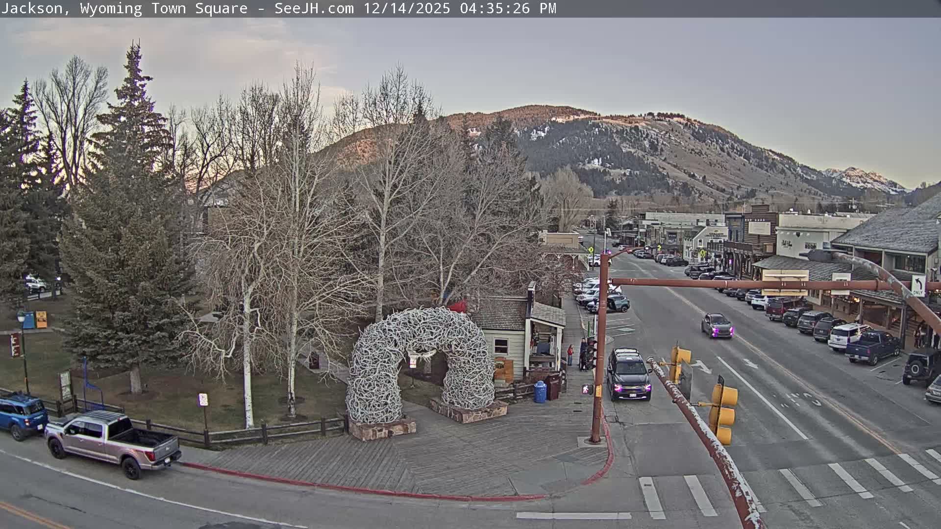 A snow-covered street and park area featuring a prominent antler archway are illuminated by artificial lights on a dark, snowy night, flanked by bare trees and buildings.