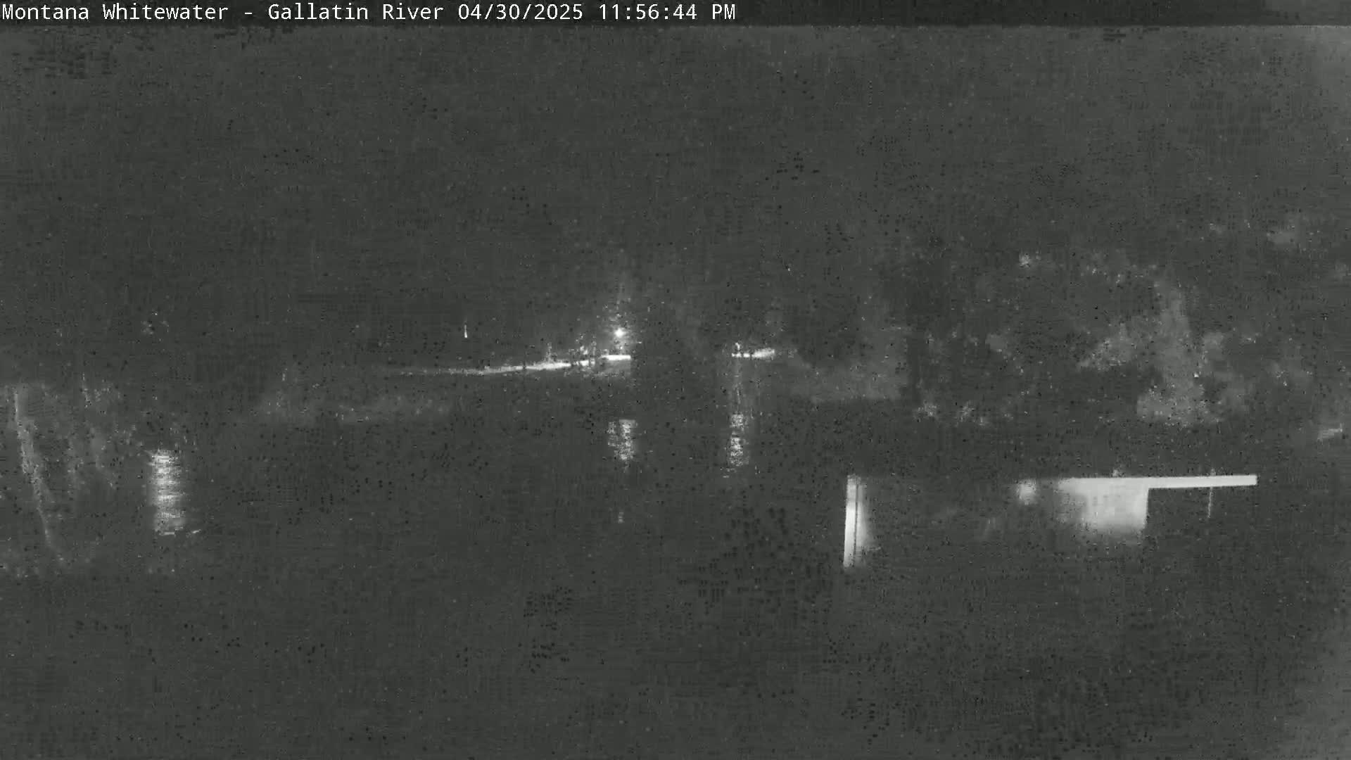 A nighttime, low-light view of a dark river reflecting a few lights, with dark vegetation visible on either side.