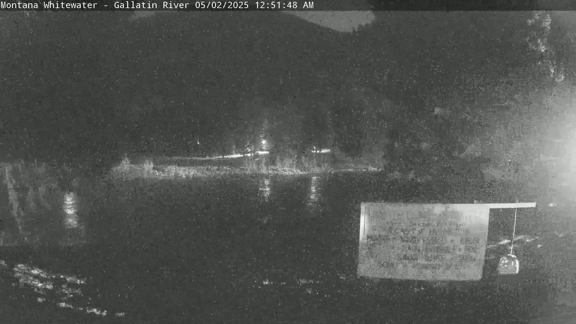 A nighttime, low-light image shows a dark body of water reflecting some lights in the distance, with dark trees lining the far bank.