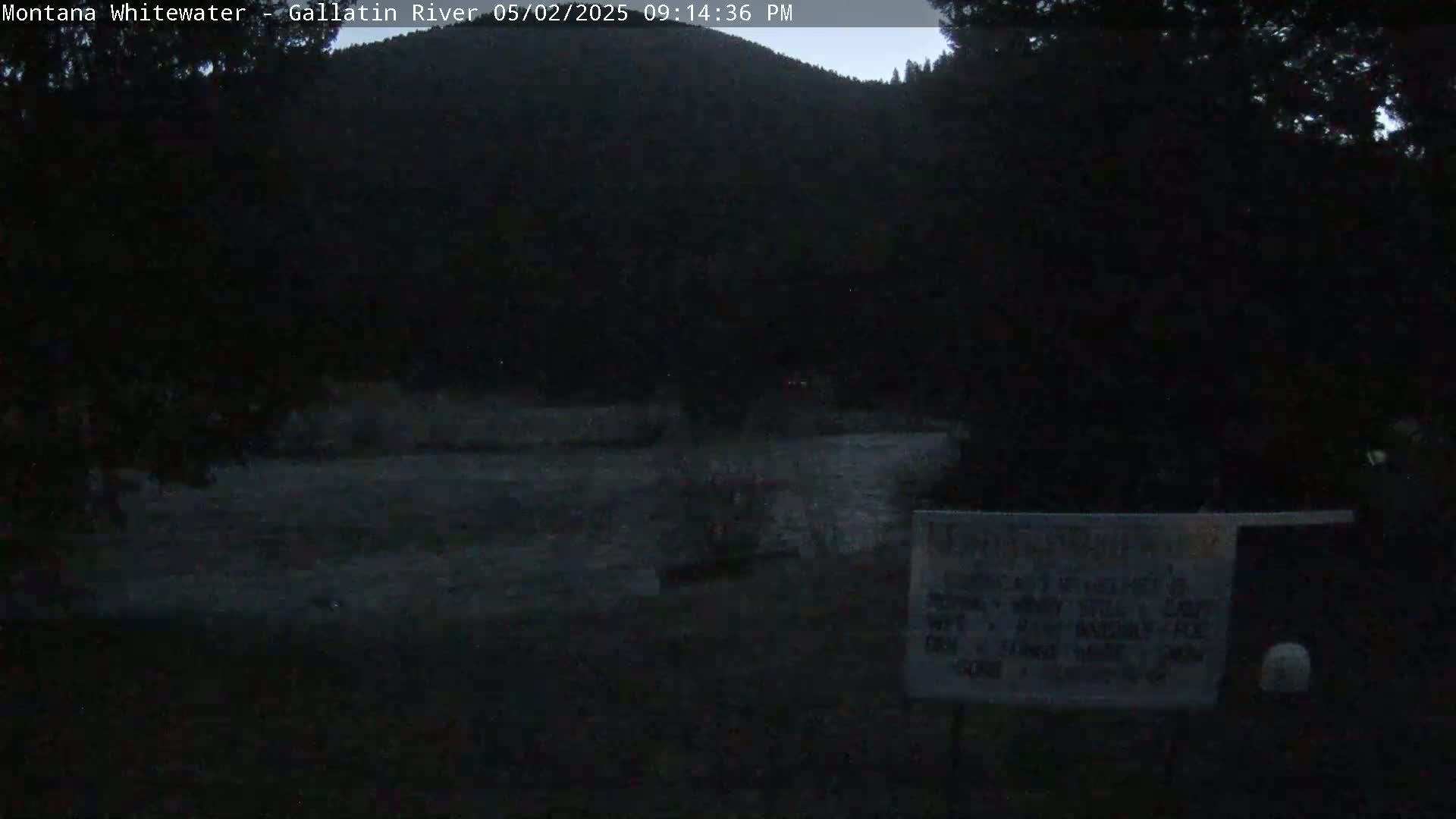 A dark, nighttime view of a river flowing past a wooded hillside under a mostly clear sky.