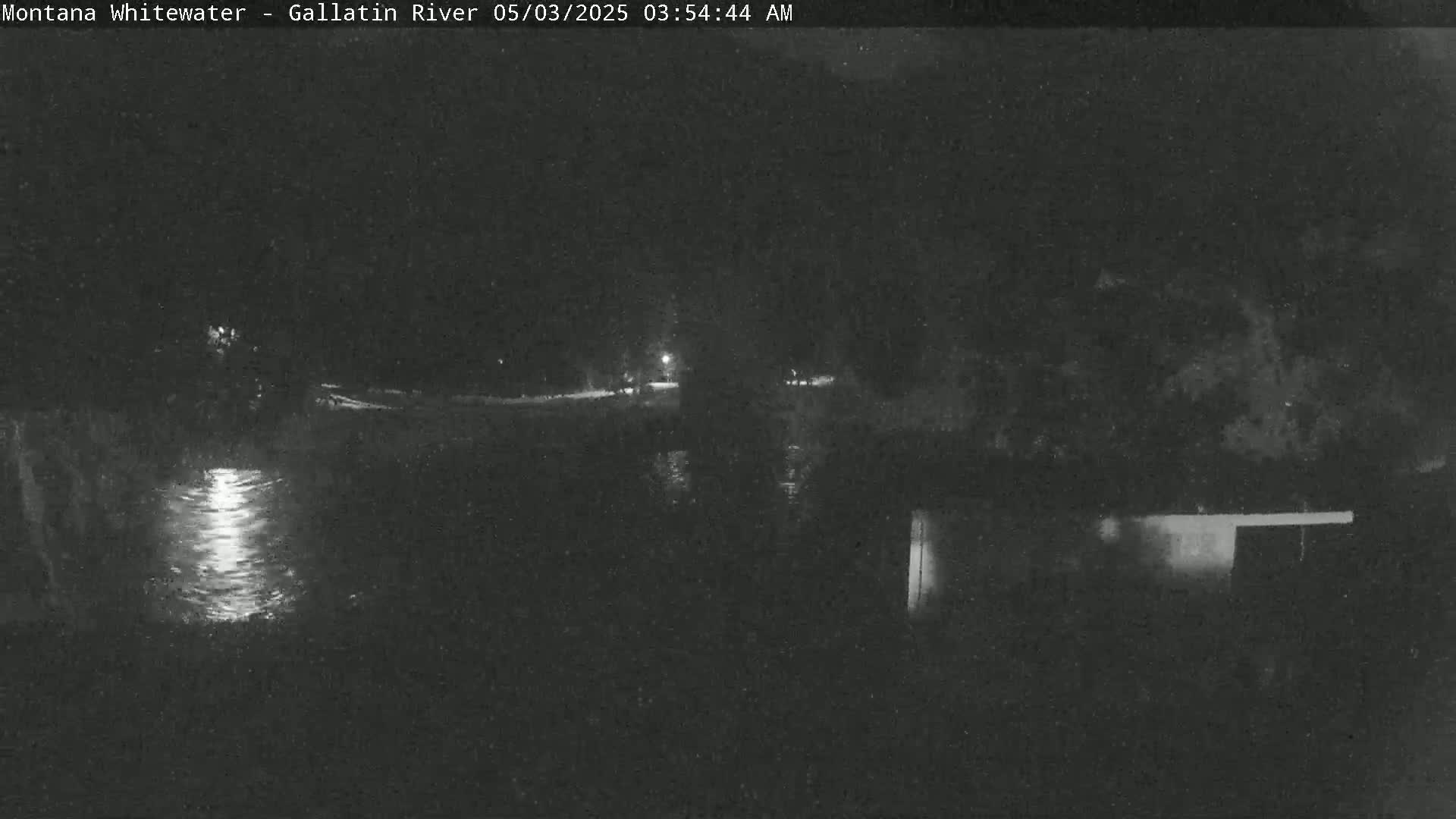 A dark, nighttime view of a river reflecting light, with trees and a building visible in the background.
