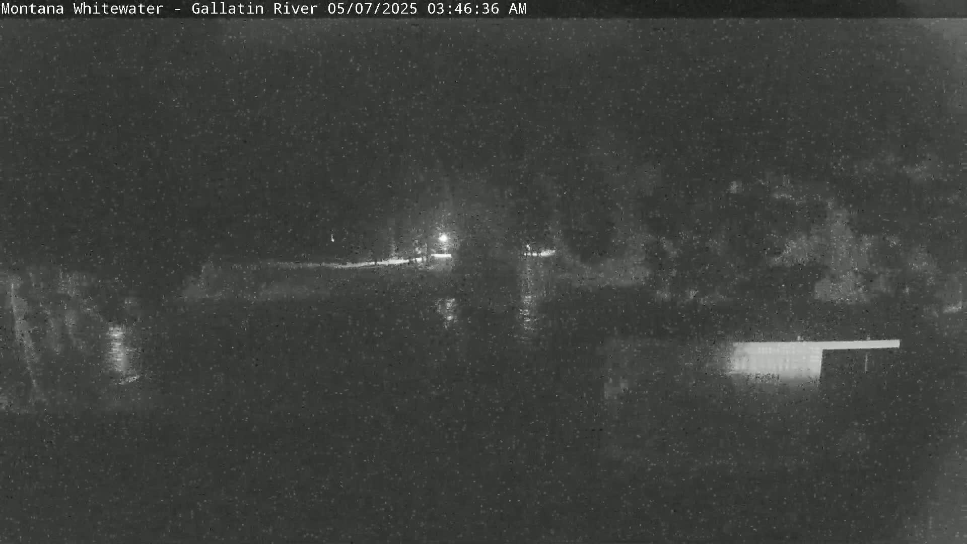 A nighttime, low-light image shows a dark body of water reflecting light, with dark vegetation on either side and a structure in the lower right corner.