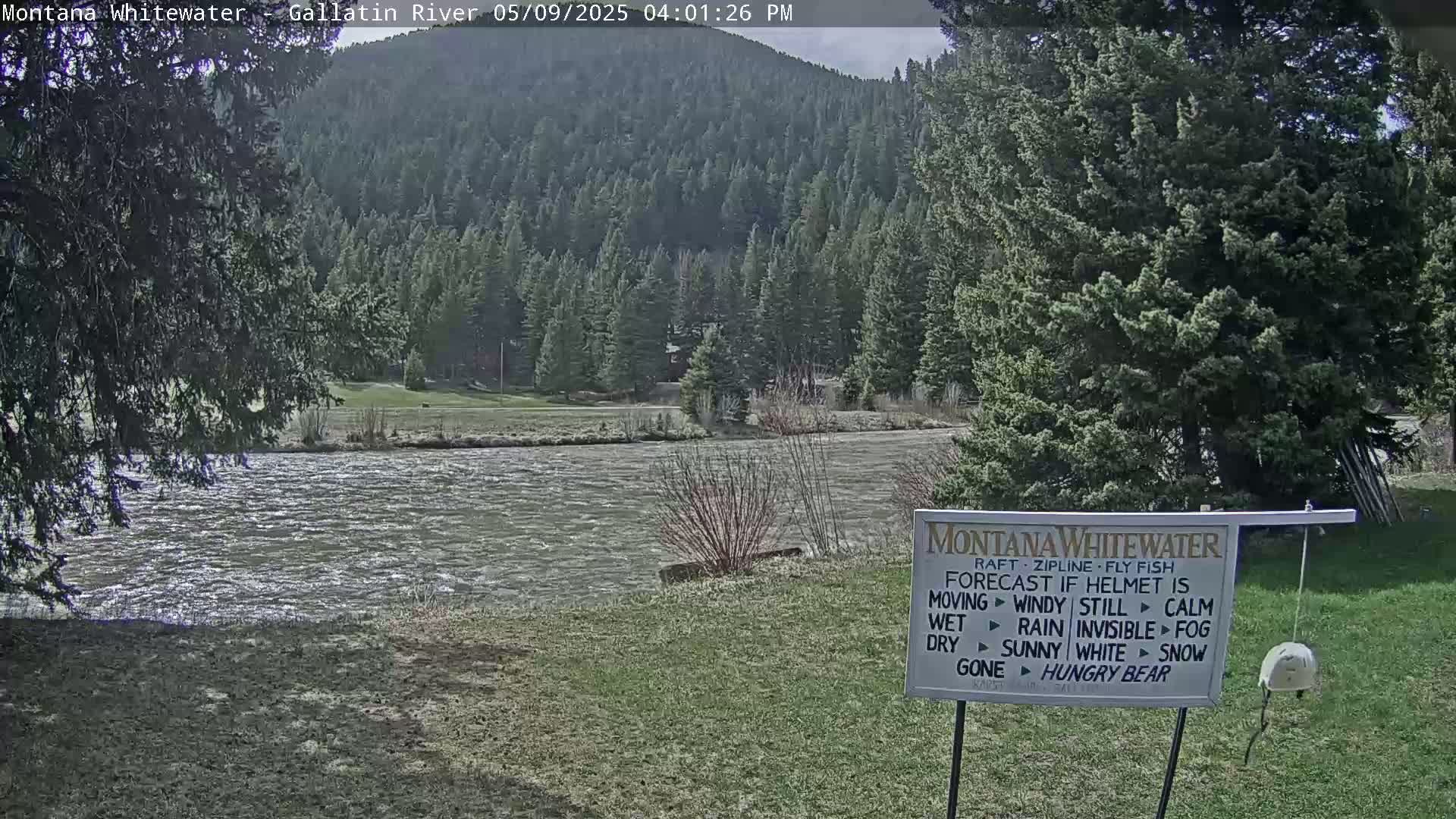 A swiftly flowing river is situated before a lush green hillside covered in evergreen trees under a partly cloudy sky.