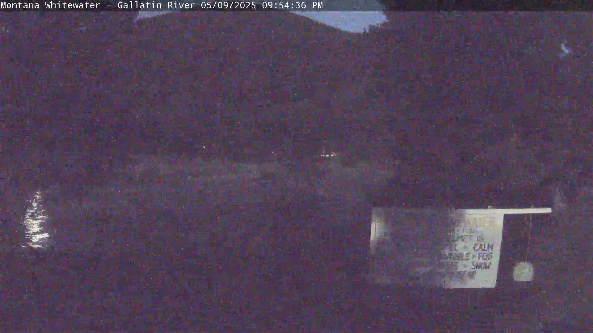 A dark, nighttime scene shows a body of water reflecting light, with a hill or mountain in the background under a calm, possibly foggy sky.