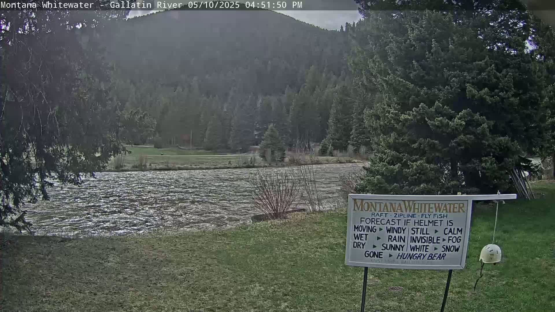 A choppy river flows in front of a grassy area and forested hills under an overcast sky.