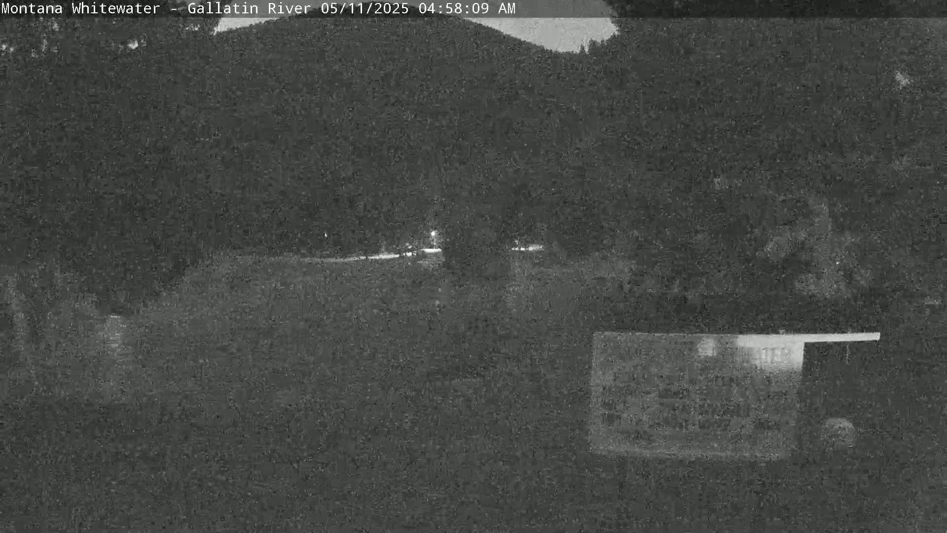 A nighttime, low-light view shows a dark, flat area in front of a wooded hillside.