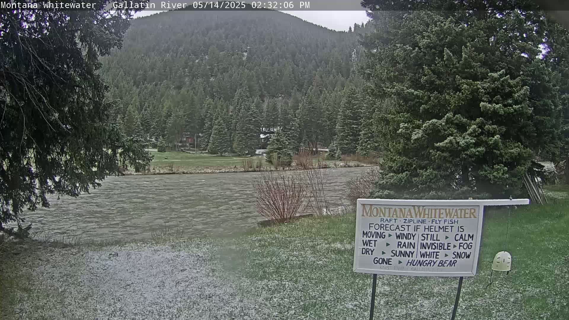 A lightly snow-covered grassy area overlooks a river flowing past a snow-dusted evergreen forest under an overcast sky.