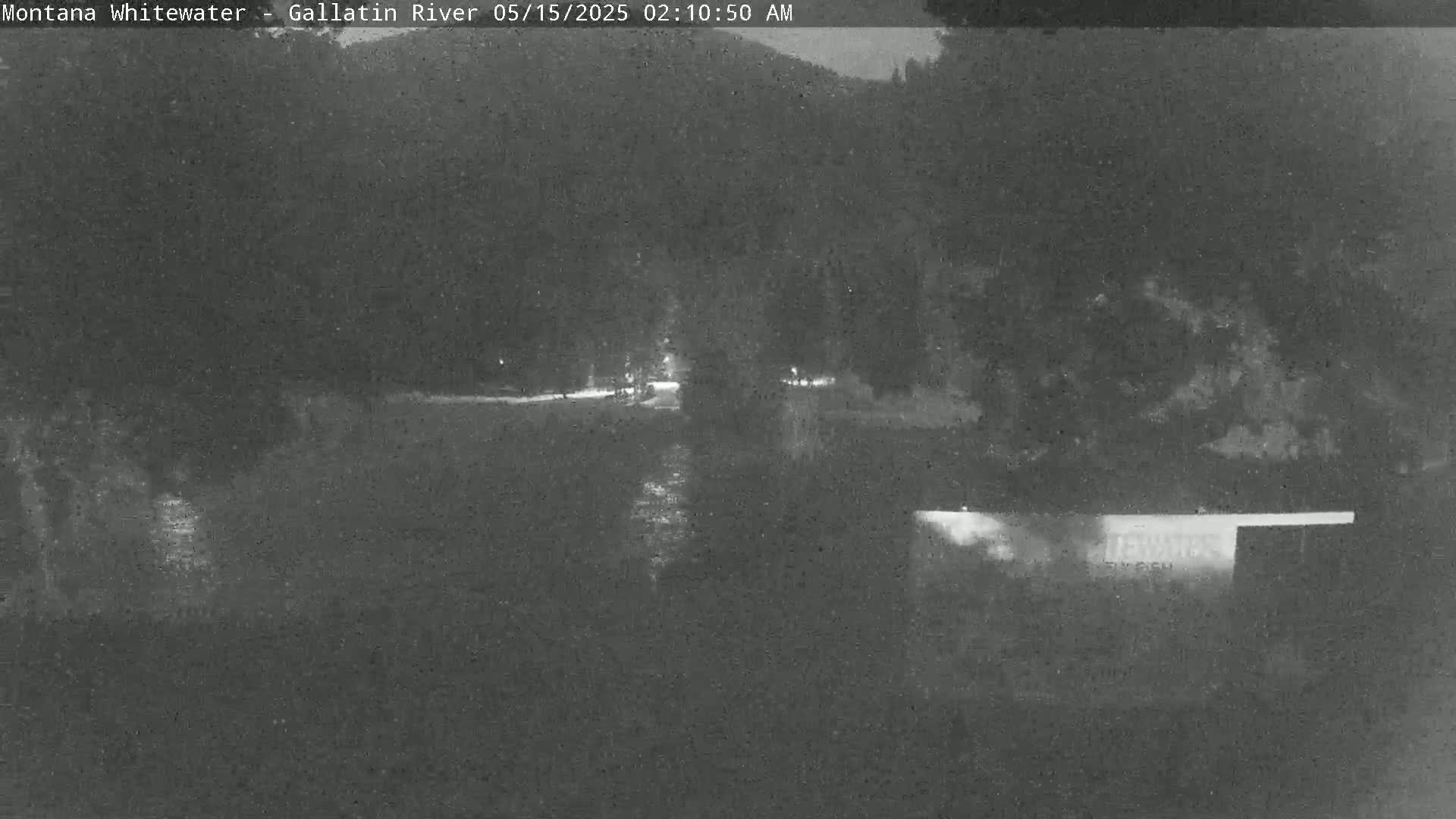 A nighttime, low-light image shows a dark river reflecting a few lights, with dark trees and land in the background under a clear, dark sky.