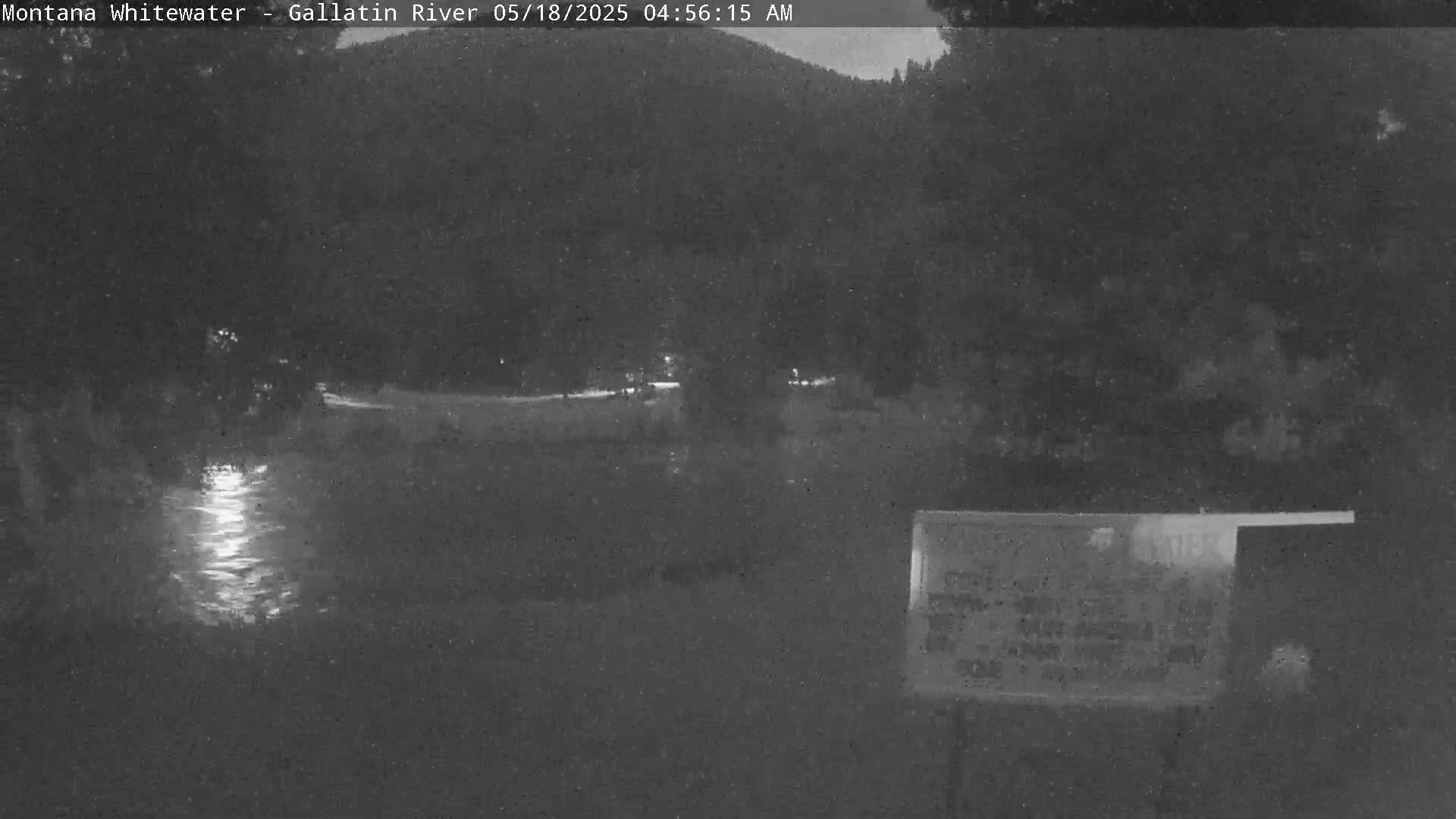 A nighttime, low-light view of a river reflecting light, with dark trees and a sign in the foreground.
