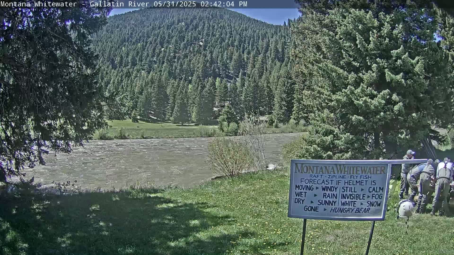 A fast-moving river flows beneath a sunny sky past a grassy bank, with evergreen-covered hills in the background and several people standing near a sign by the river's edge.